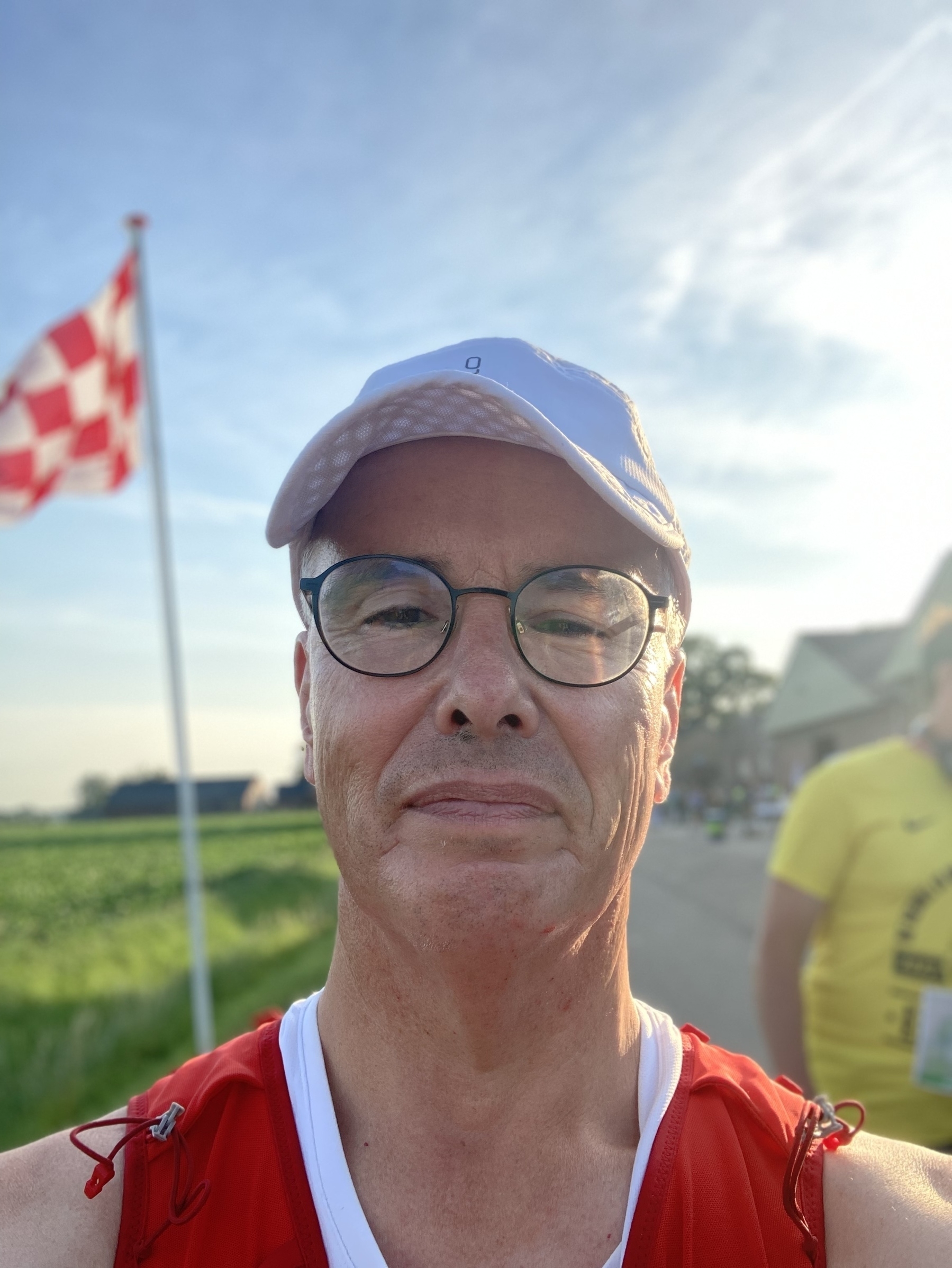 Een persoon met een bril en een pet staat buiten in de buurt van een geruite vlag, met een heldere lucht en andere mensen op de achtergrond.