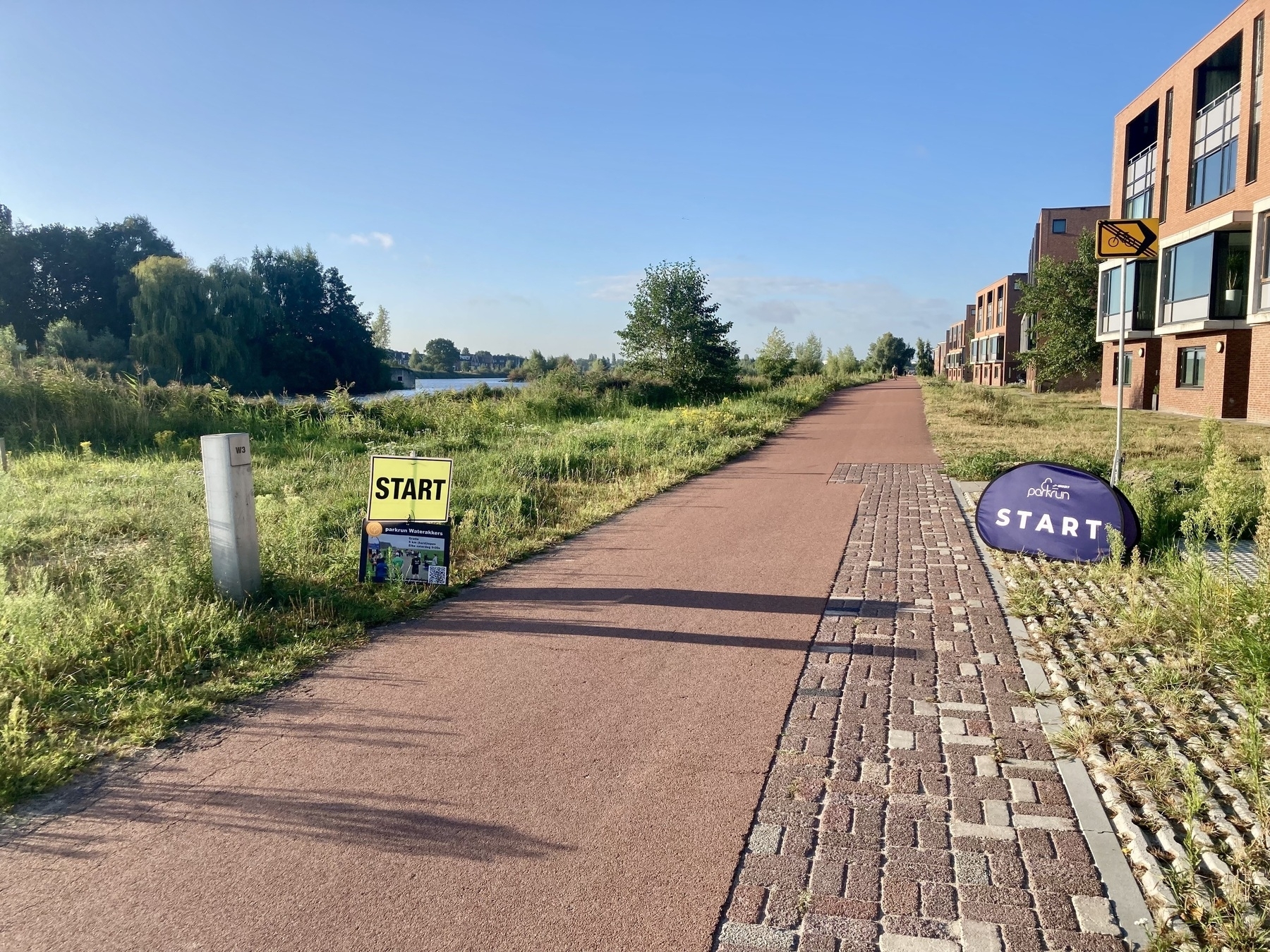 Een pad loopt tussen een grasveld en enkele gebouwen, met borden die een startpunt aangeven voor een evenement of activiteit.