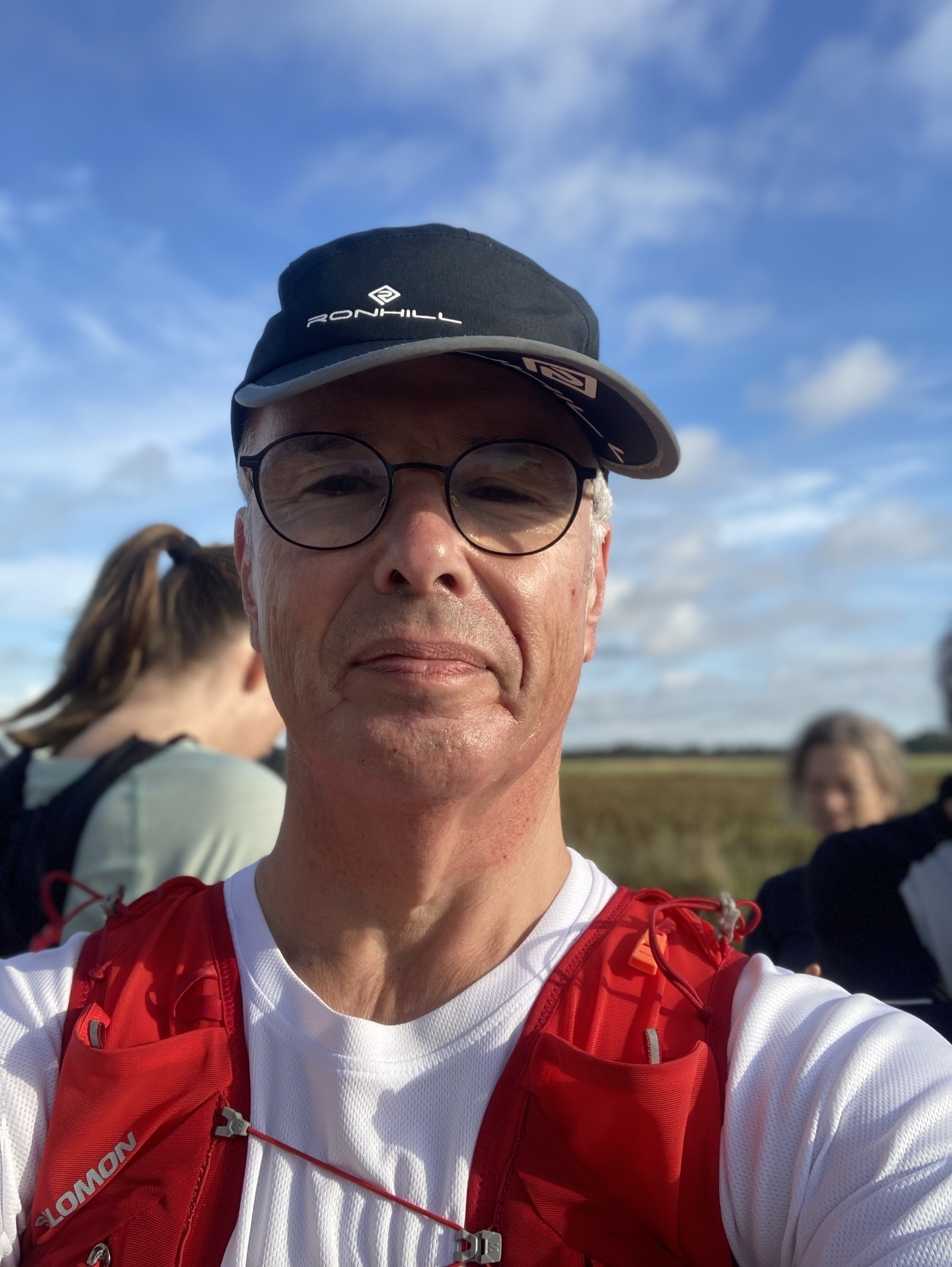 Een persoon met een pet en bril is buiten, vergezeld door andere mensen op de achtergrond.