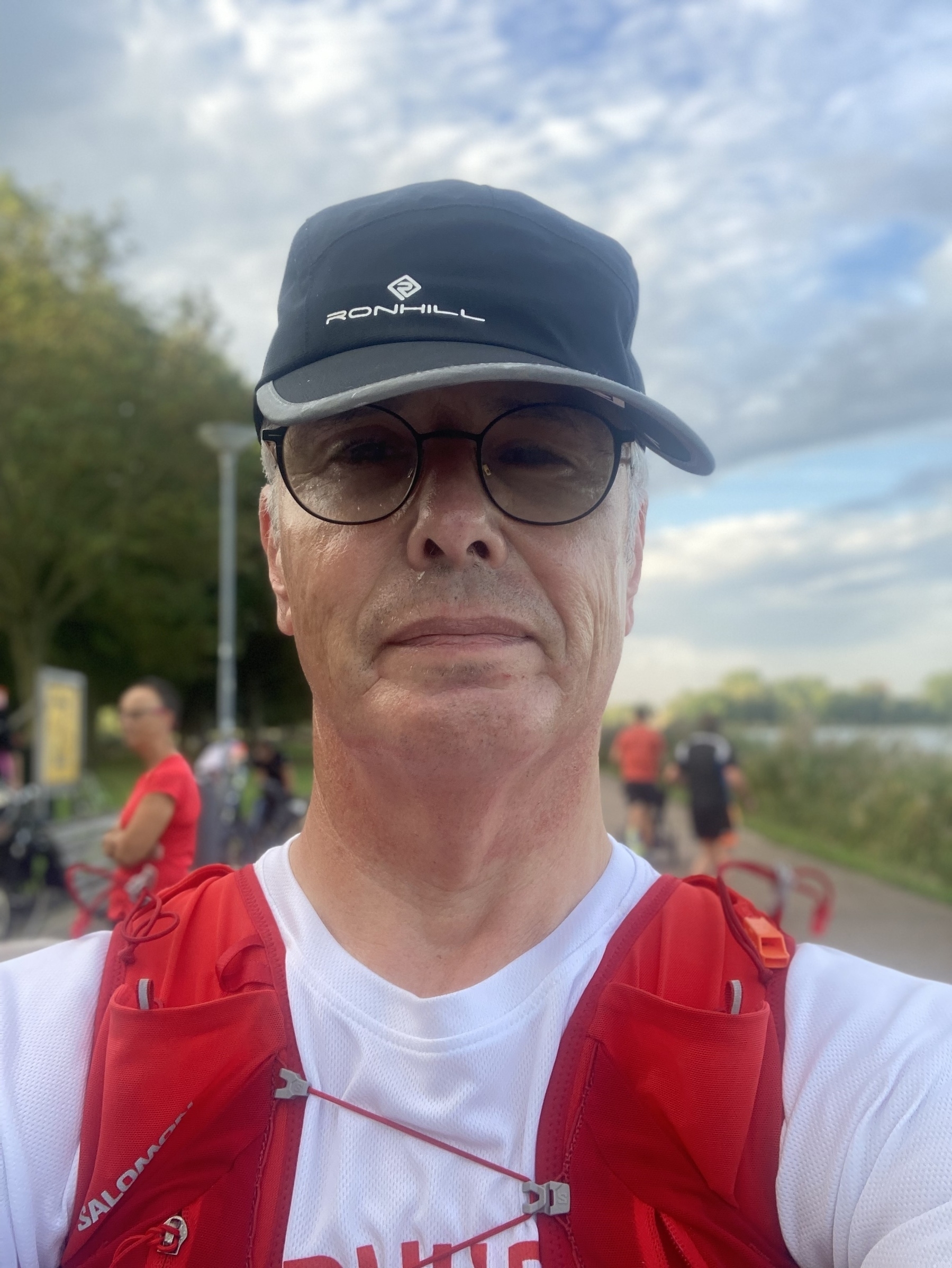 Een man met een bril, een baseballpet en een rood vest is buiten op een pad in de buurt van het groen en andere mensen.