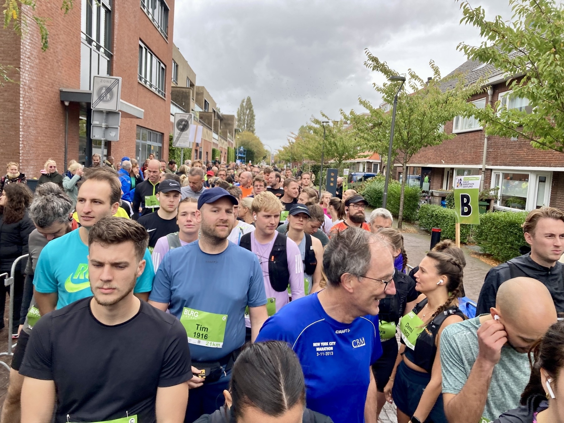Een grote groep mensen, velen dragen borstnummers, zijn buiten op straat verzameld, ter voorbereiding op een hardloopevenement.