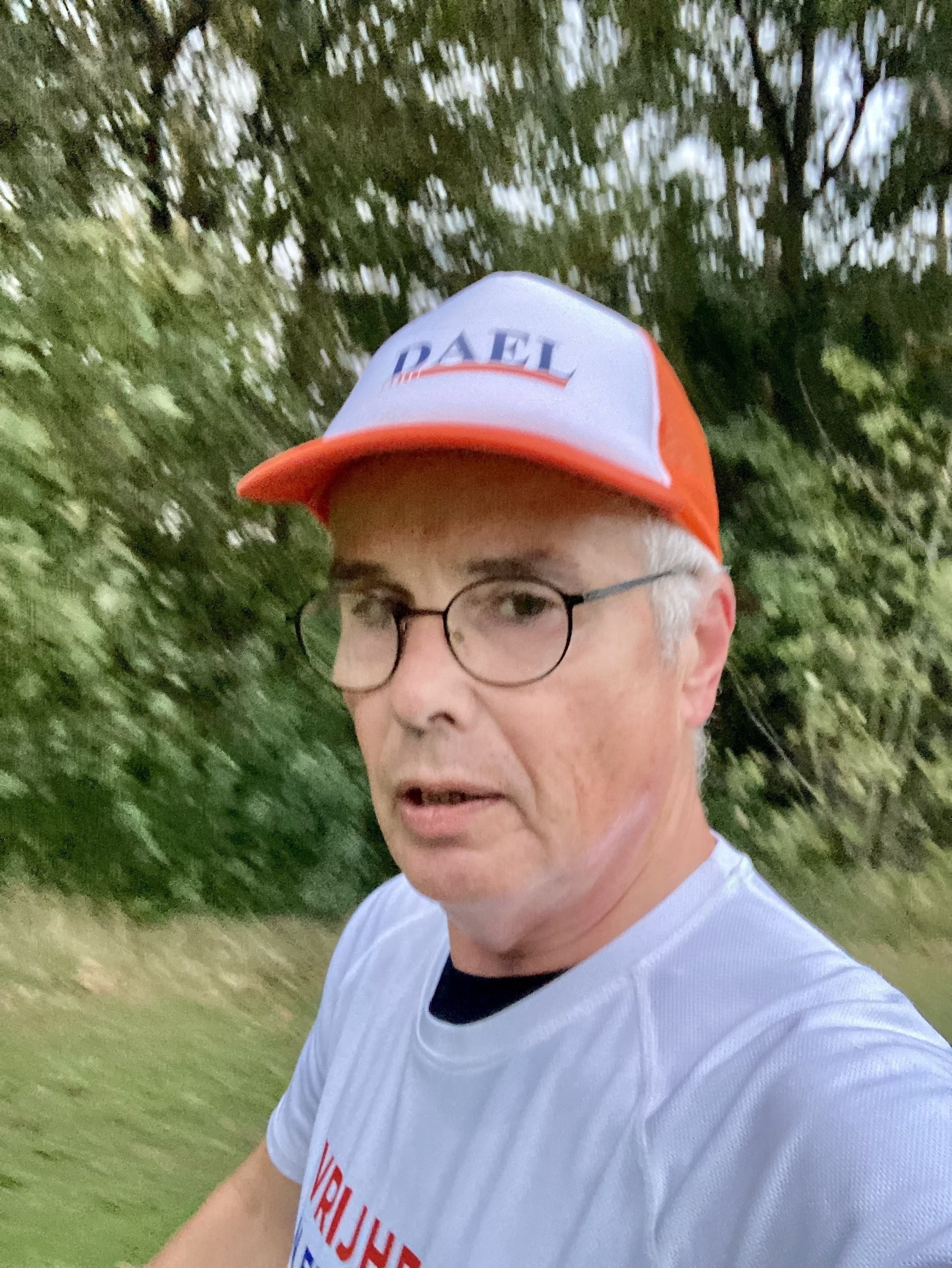 Een persoon met een bril, een rood-witte pet en een wit shirt is buiten, schijnbaar bezig met een fysieke activiteit zoals joggen, met groen op de achtergrond.