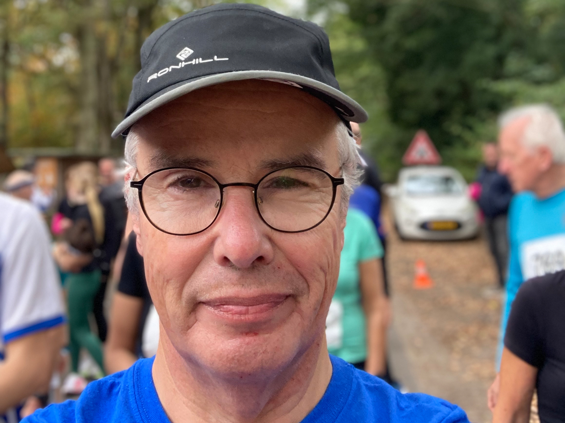 Een man met een hoed en een bril is buiten met mensen en bomen op de achtergrond.