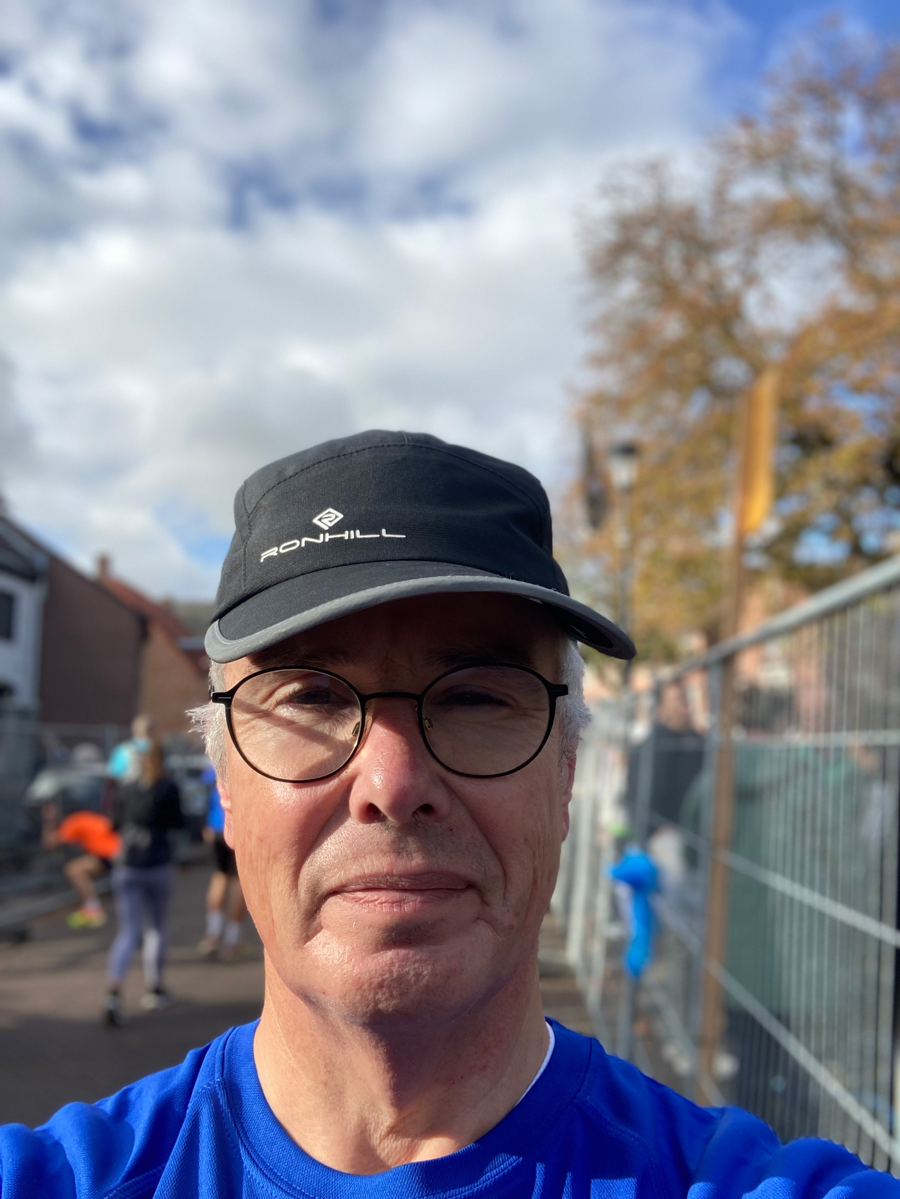 Een persoon met een blauw shirt en een zwarte pet neemt een selfie buiten, met mensen en gebouwen op de achtergrond.