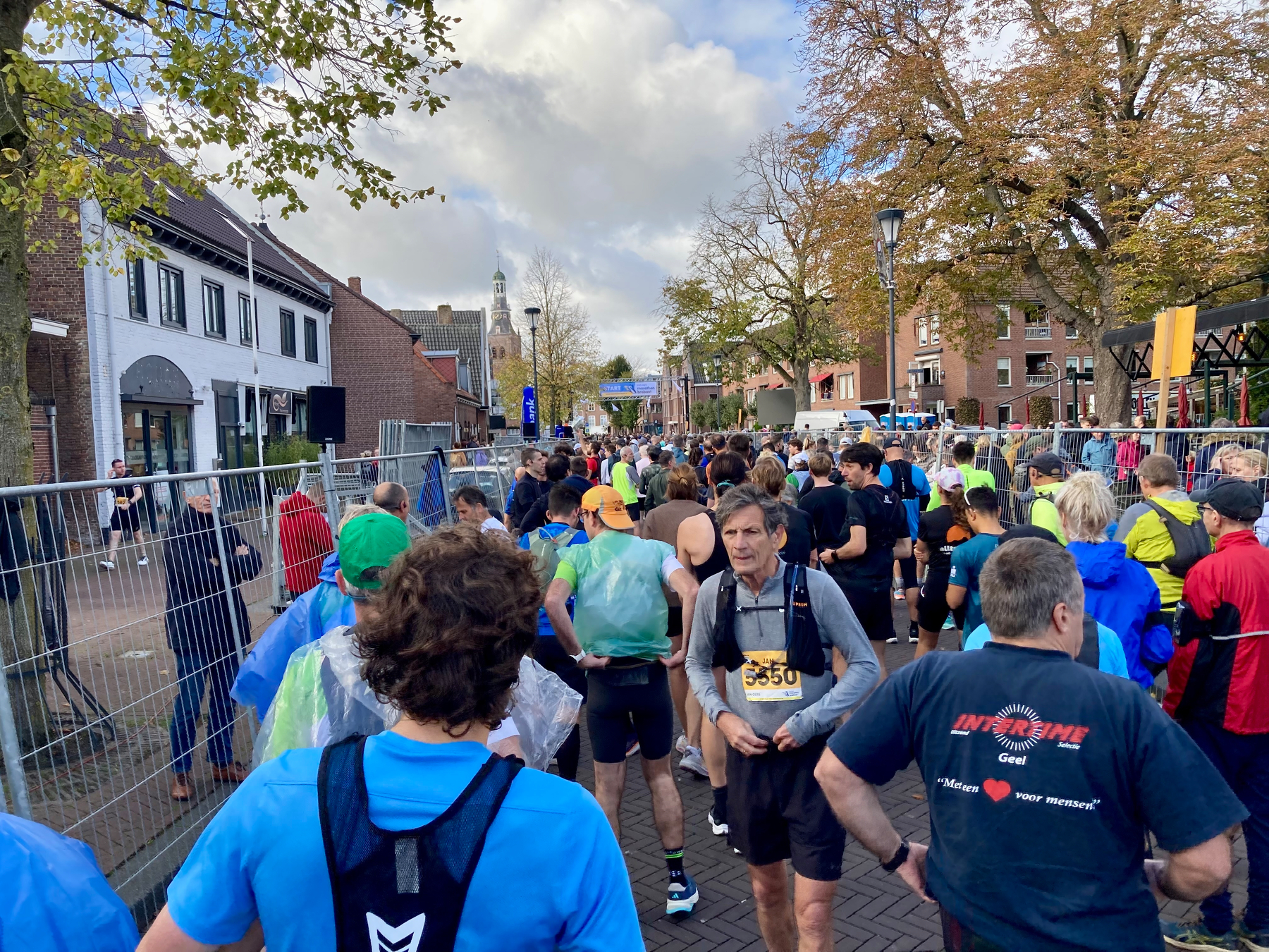 Een groep mensen met hardloopuitrusting verzamelt zich bij de startlijn van een wedstrijd in een straat omzoomd met bomen en gebouwen.