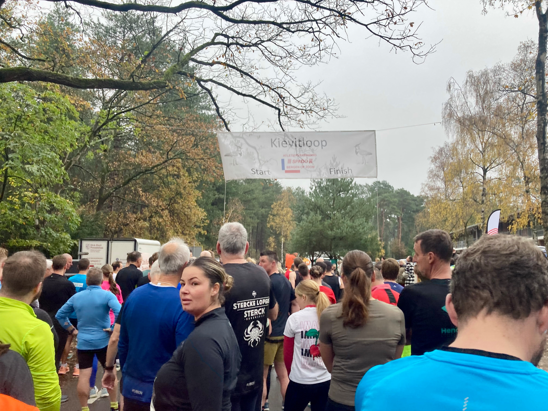 Een groep mensen verzamelt zich aan de startlijn van een hardloopevenement in een park met herfstbomen.