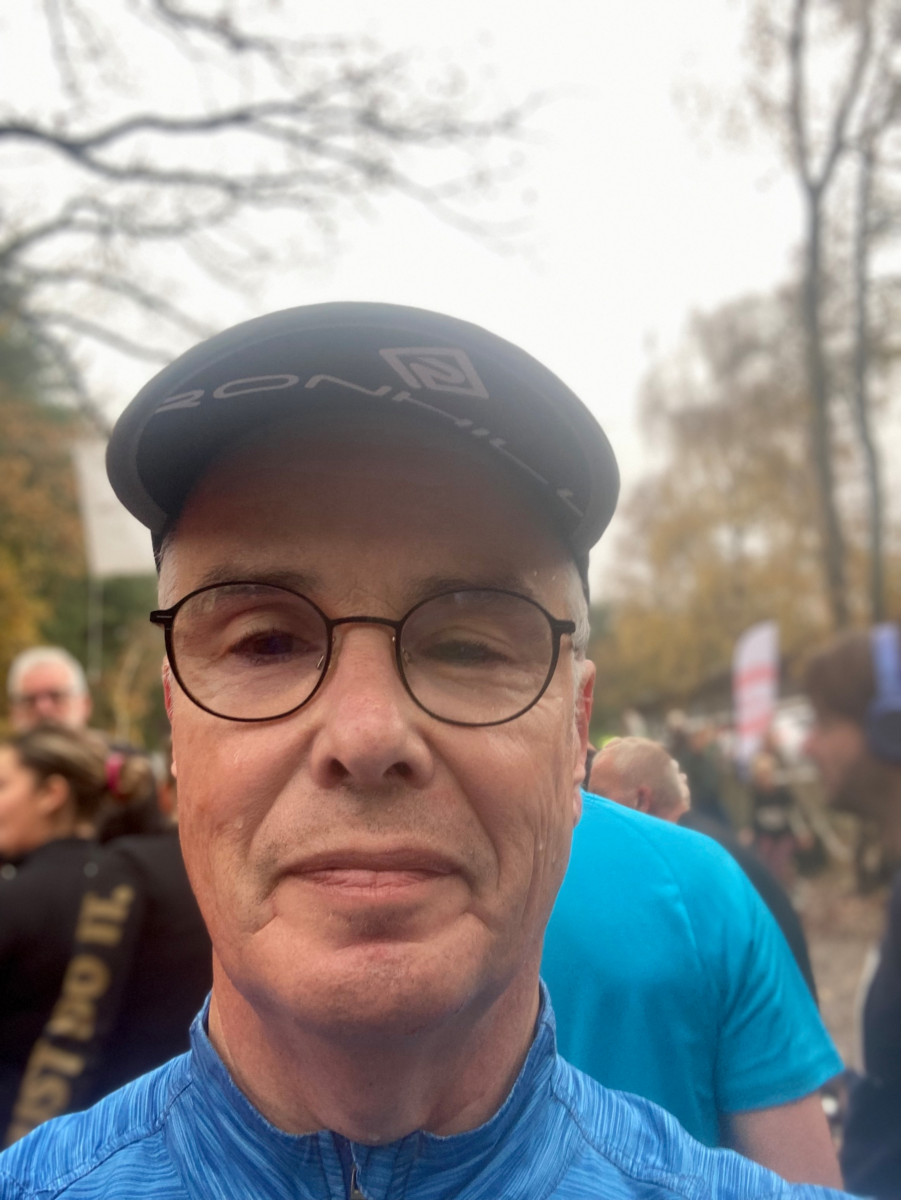 Een persoon met een bril en een pet is in een groepsomgeving buiten, omringd door andere mensen.