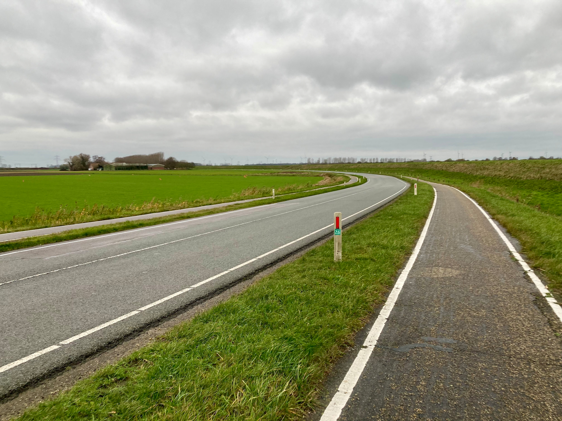 Een kronkelende weg en aangrenzende fietspad strekken zich uit door vlakke groene velden onder een bewolkte lucht.