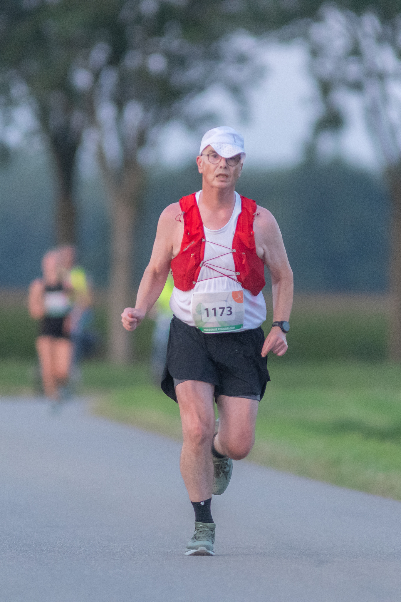 Een persoon rent tijdens een race op een weg, draagt een witte pet, een rood vest en een borstnummer, met wazige bomen en andere lopers op de achtergrond.