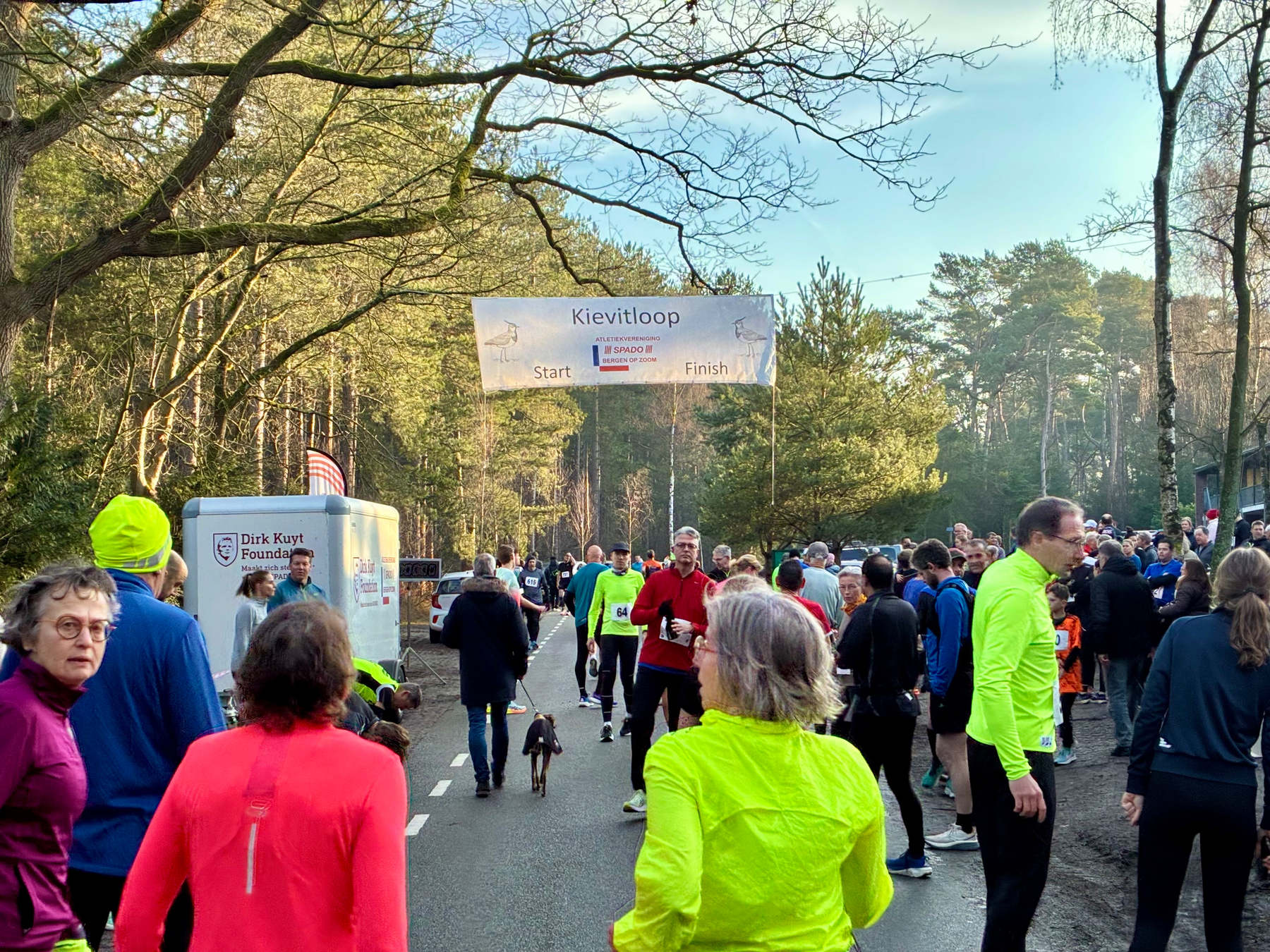 Een groep mensen, sommigen met kleurrijke atletische uitrusting, nemen deel aan een buitenevenement gemarkeerd met een Kievitloop start- en finishbanner.