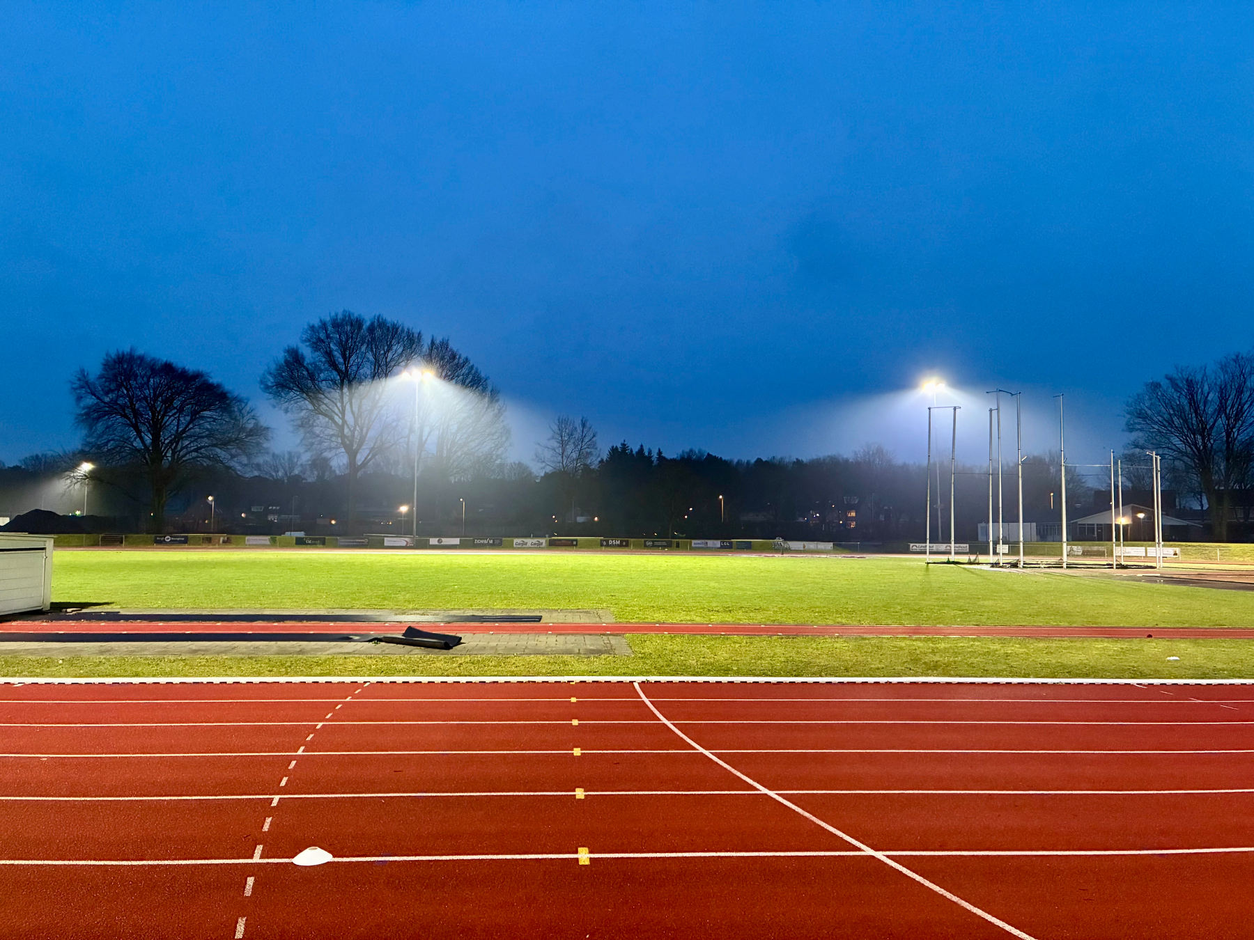 Een goed verlicht sportveld met een hardloopbaan is in de vroege avond omringd door bomen.