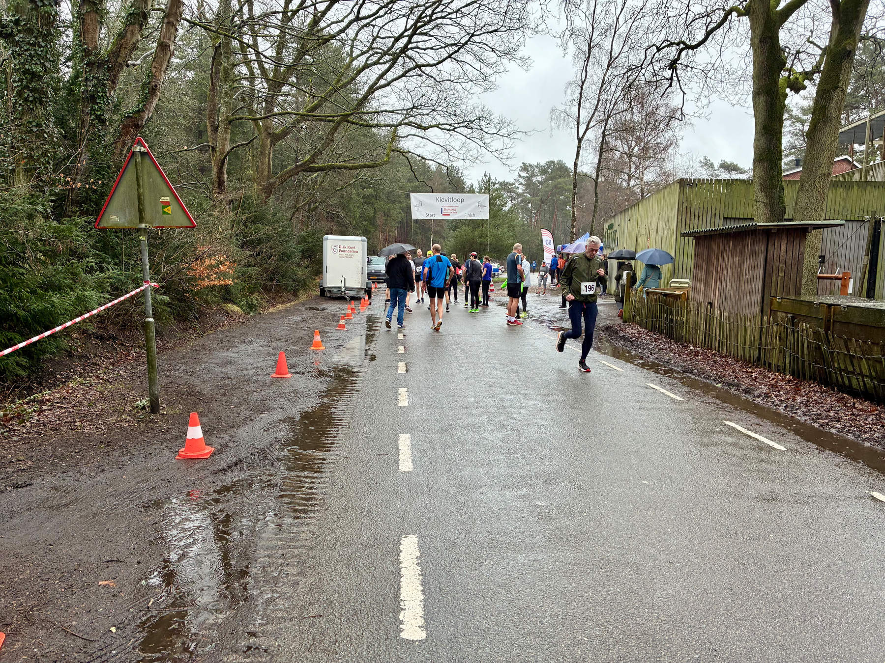 Een groep hardlopers neemt deel aan een race langs een natte weg omringd door bomen.