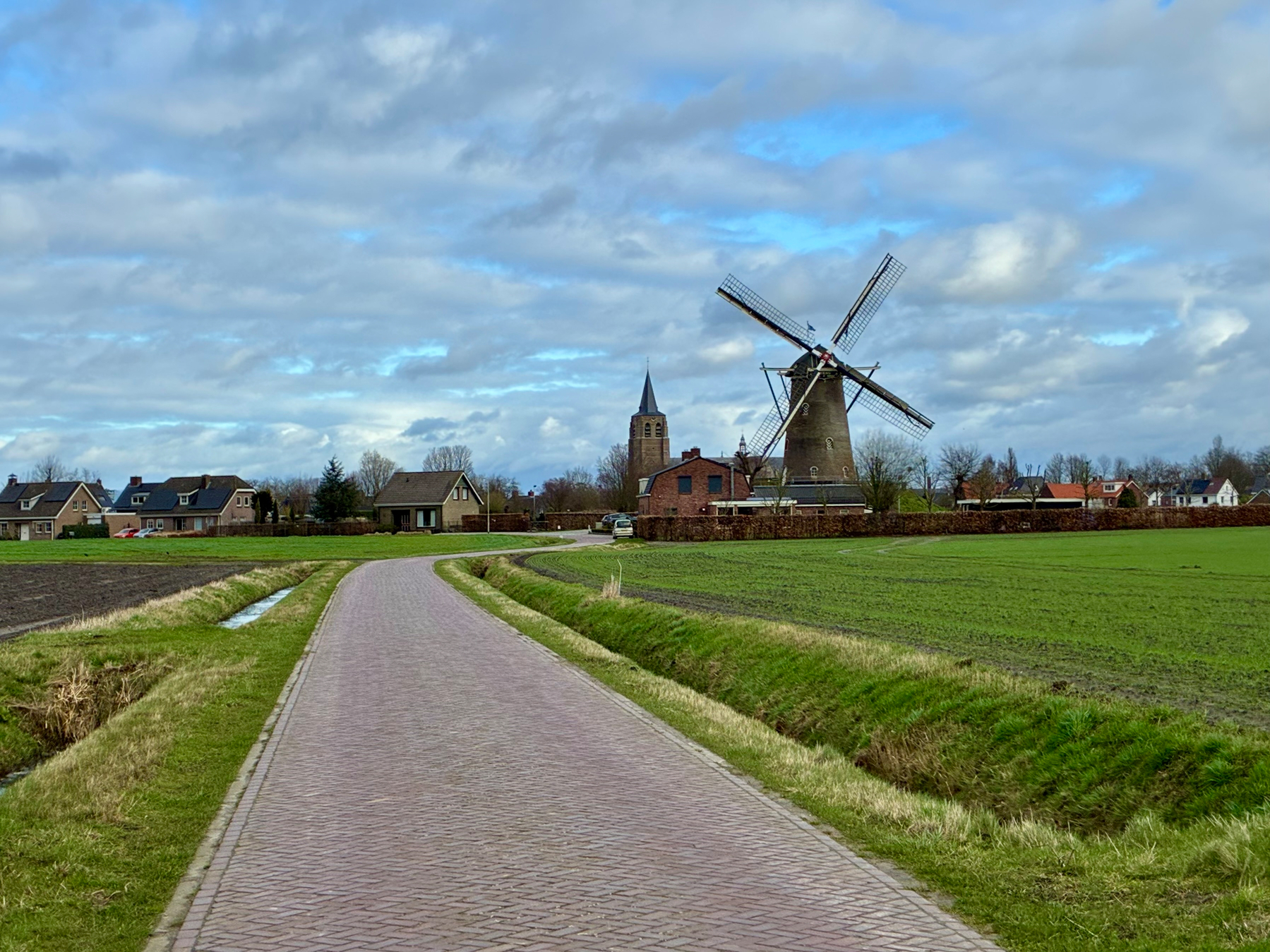 Een landschapsscène heeft een kronkelend geplaveid pad dat leidt naar een traditionele windmolen, omringd door velden en een paar huizen onder een gedeeltelijk bewolkte lucht.