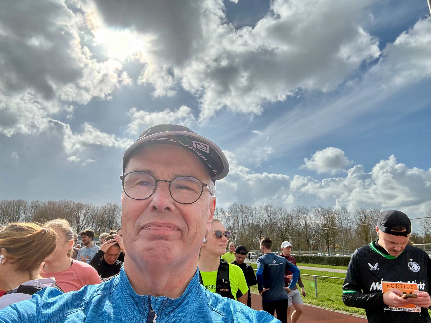 Een groep mensen, waaronder een man met een bril en een pet die een selfie maakt, wordt buiten verzameld op een zonnige en deels bewolkte dag.