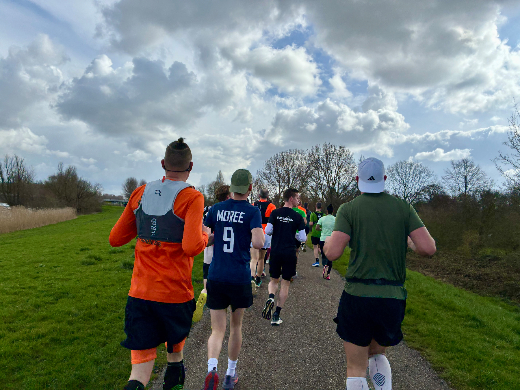 Een groep mensen rent samen op een verhard pad omringd door grasvelden en bomen onder een bewolkte lucht.