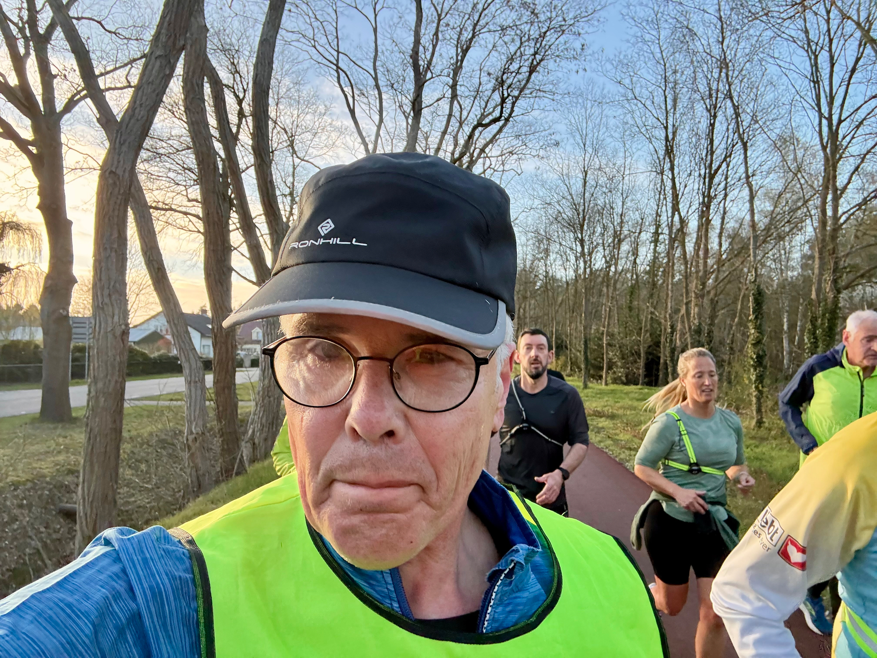 Een groep mensen, waaronder een man in een geel vest en pet, jogt op een pad omringd door bomen.