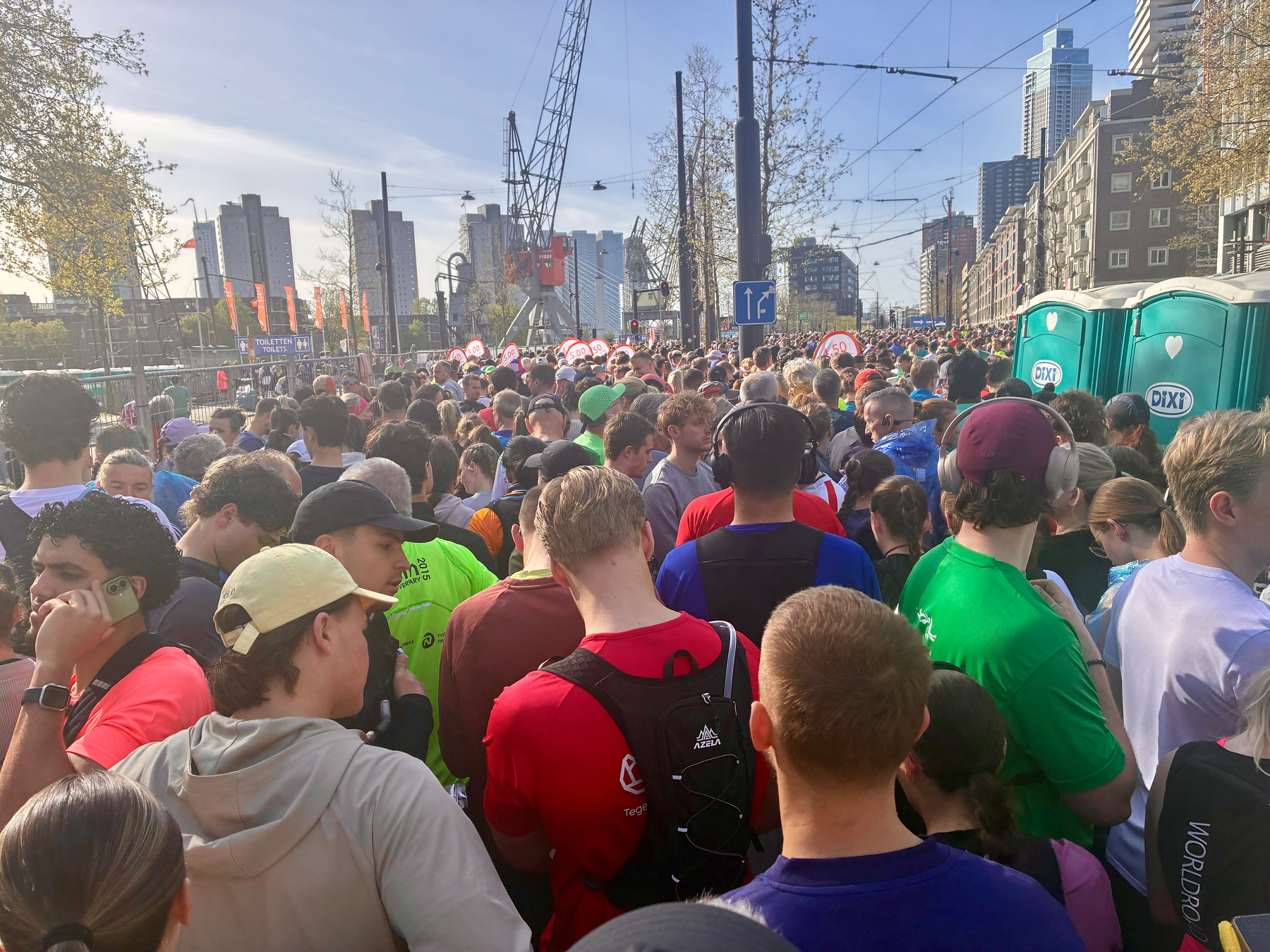 Een grote menigte mensen, velen in hardloopkleding, zijn verzameld in de buurt van draagbare toiletten en gebouwen op de achtergrond onder een heldere lucht.