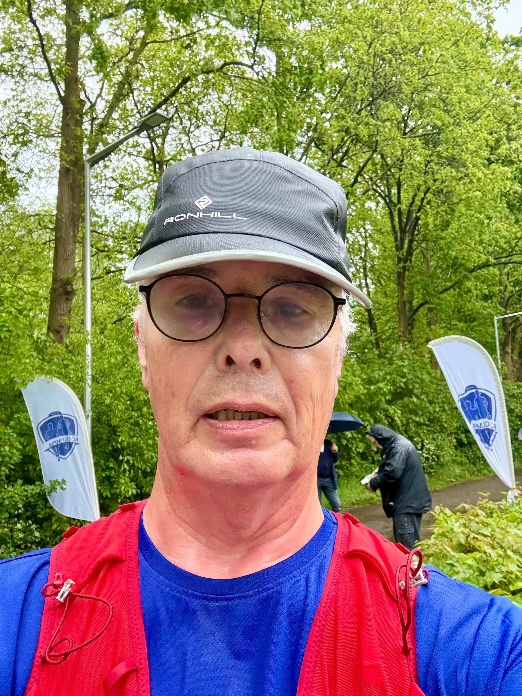 Een persoon met een Ronhill-pet en een bril maakt een selfie buiten in de buurt van enkele spandoeken, met bomen op de achtergrond.