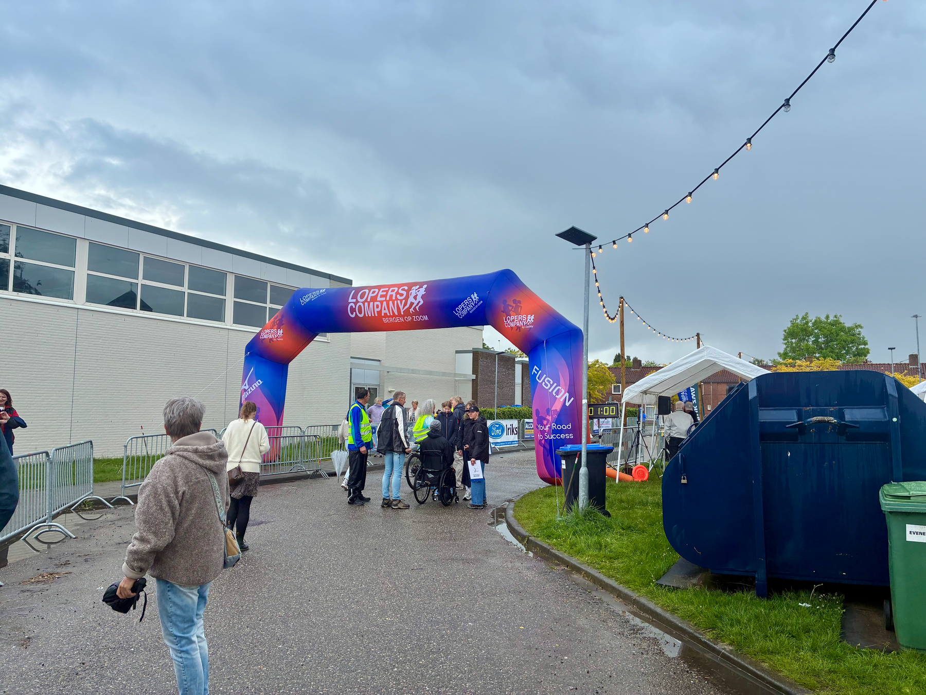Een groep mensen is verzameld onder een opblaasbare boog met evenementbranding, in de buurt van een blauwe container en buitenverlichting, op een bewolkte dag.