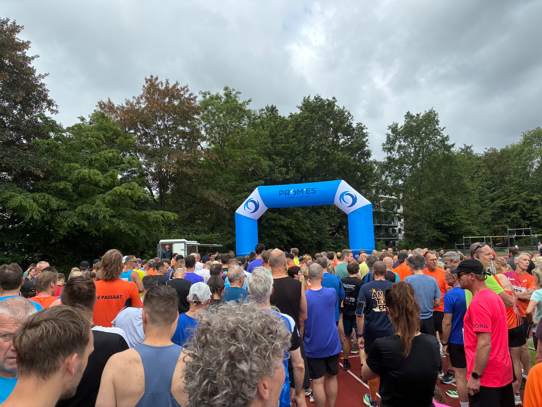 Een grote groep hardlopers is verzameld onder een blauwe start-/finish-boog tijdens een buitenevenement.