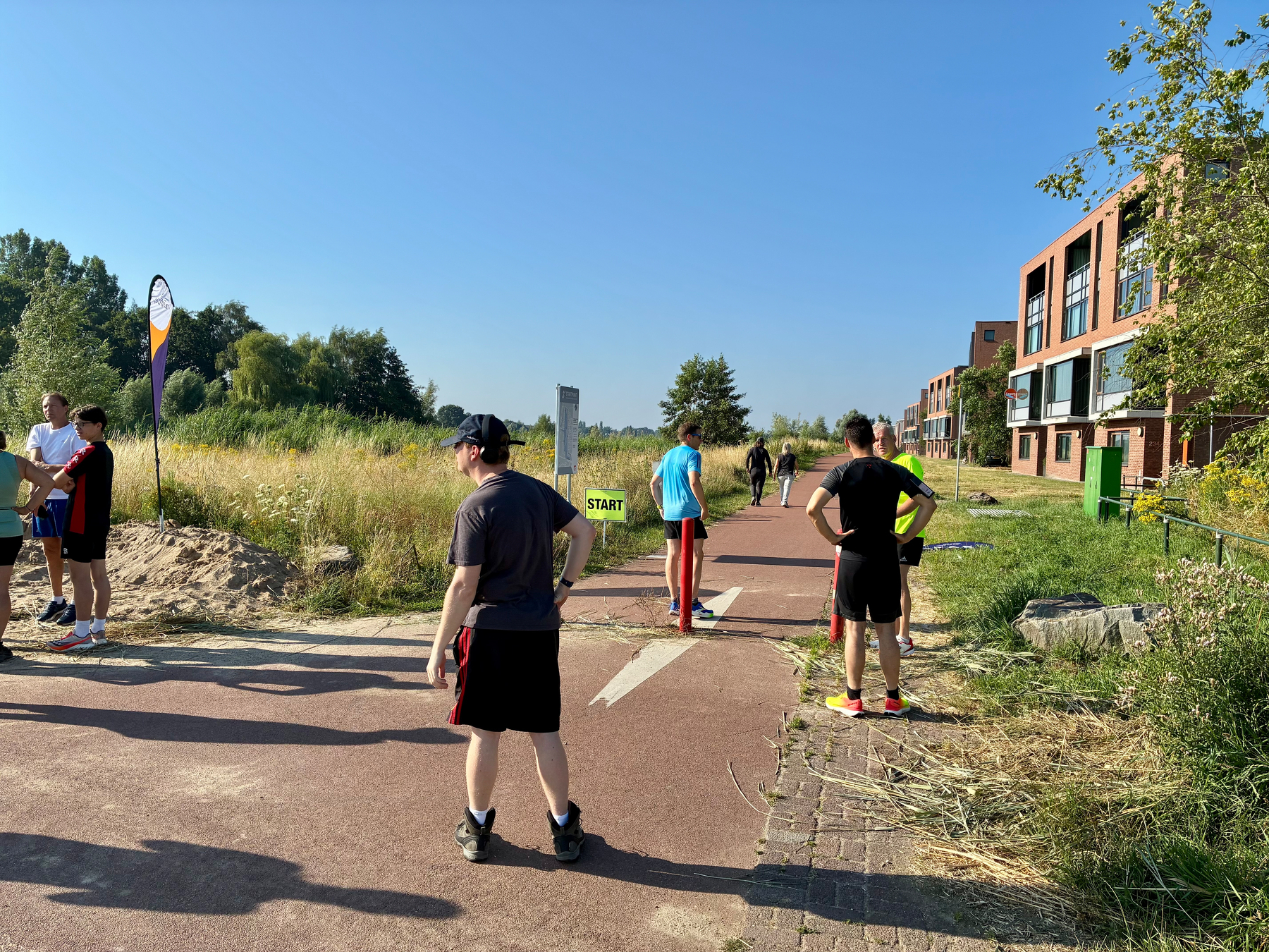 Verschillende mensen staan te wachten op een verhard pad in de buurt van een START-markering, met enkele gebouwen en groen op de achtergrond onder een heldere blauwe lucht.