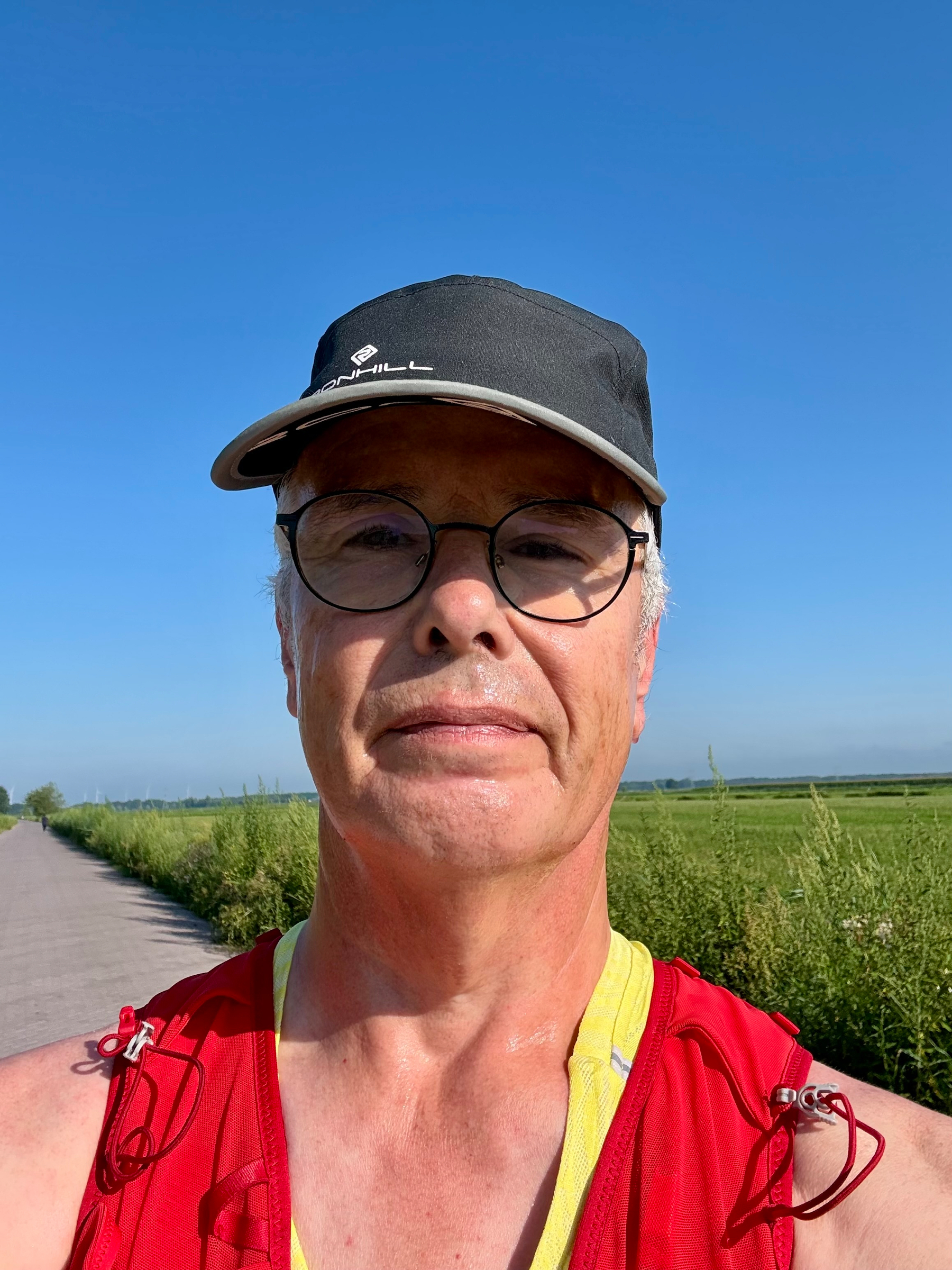 Een man met een bril, een zwarte pet en een rood vest staat op een pad met groene velden en een heldere blauwe lucht op de achtergrond.