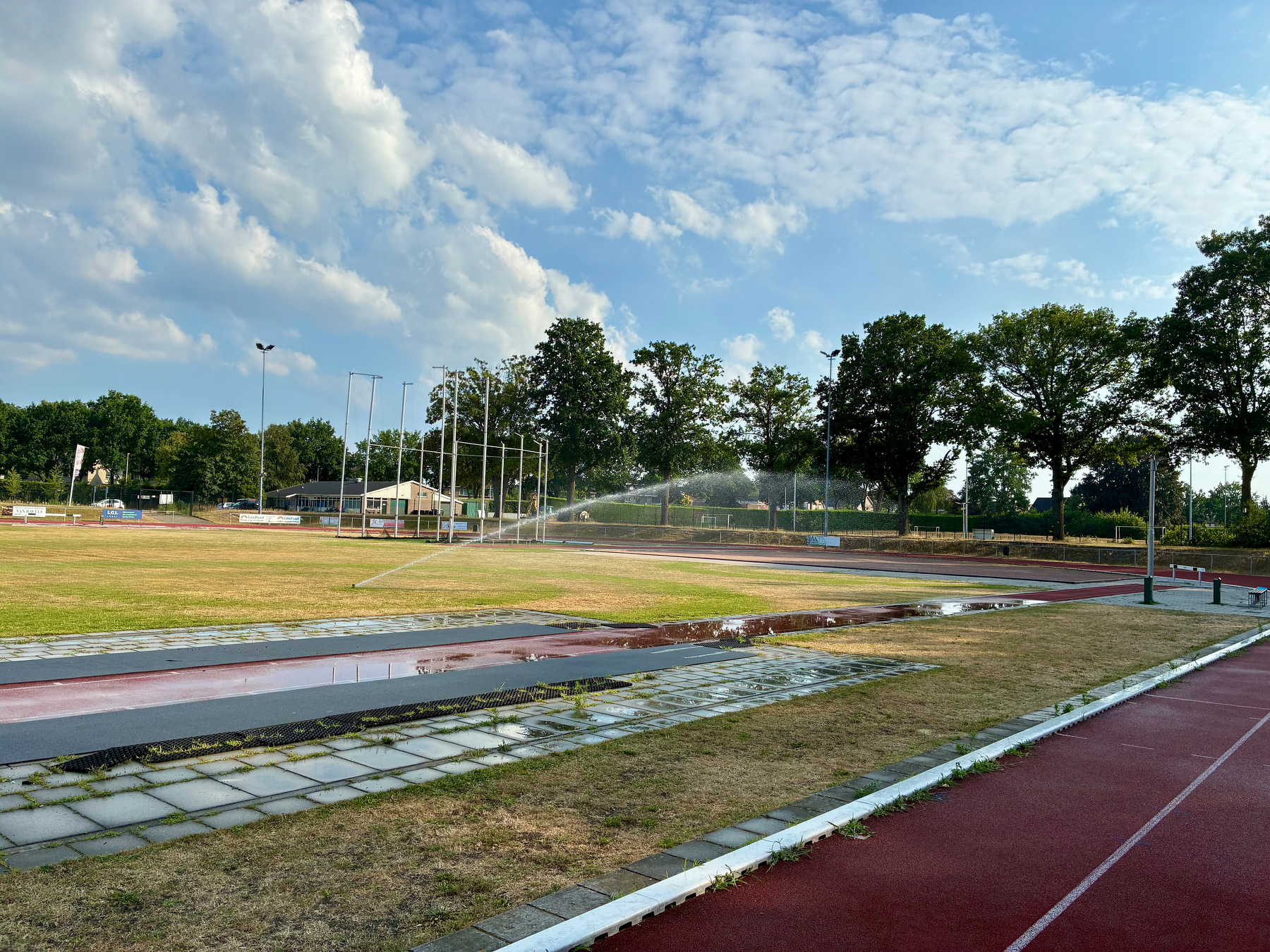 Een sportveld en een hardloopbaan zijn te zien onder een deels bewolkte hemel met sprinklers die het gras bewateren.