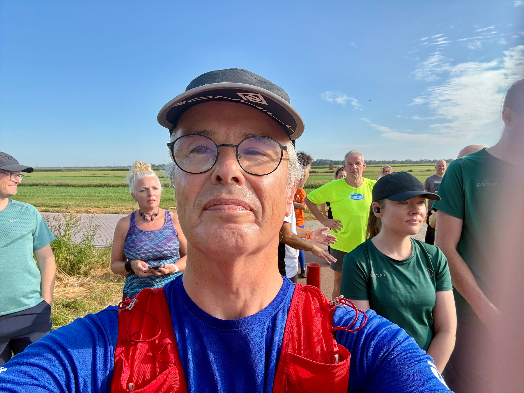 Een groep mensen gekleed in sportkleding verzamelt zich buiten op een zonnige dag, met een man met een bril en een pet die een selfie op de voorgrond maakt.