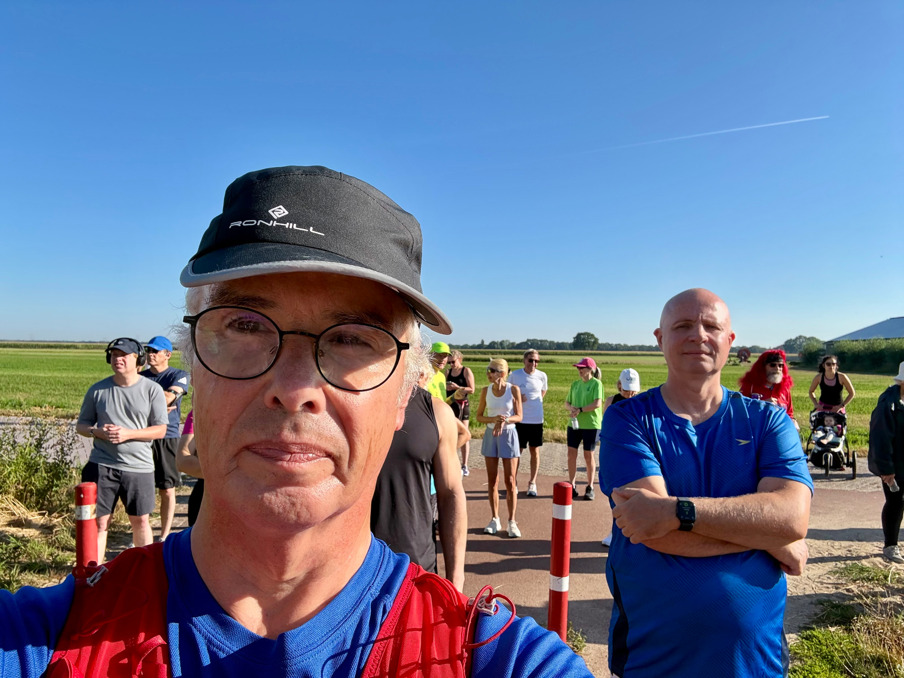 Een groep mensen, gekleed in atletische kleding, verzamelt zich buiten onder een heldere blauwe lucht, met een man met een bril en een zwarte pet die een selfie maakt op de voorgrond.