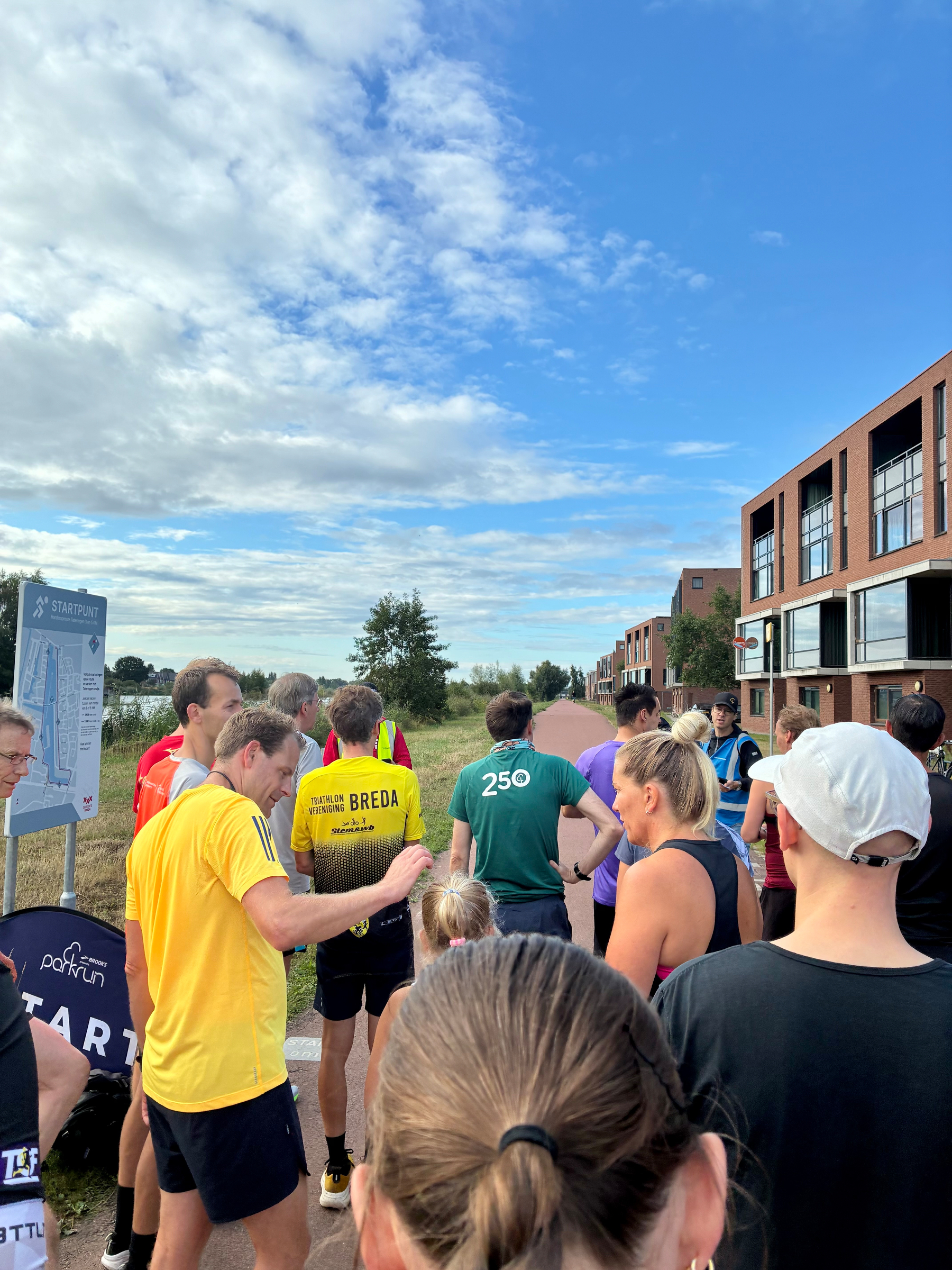 Een groep mensen verzamelt zich buiten bij een startpuntbord, om zich voor te bereiden op een parkrun op een pad naast moderne gebouwen onder een deels bewolkte lucht.