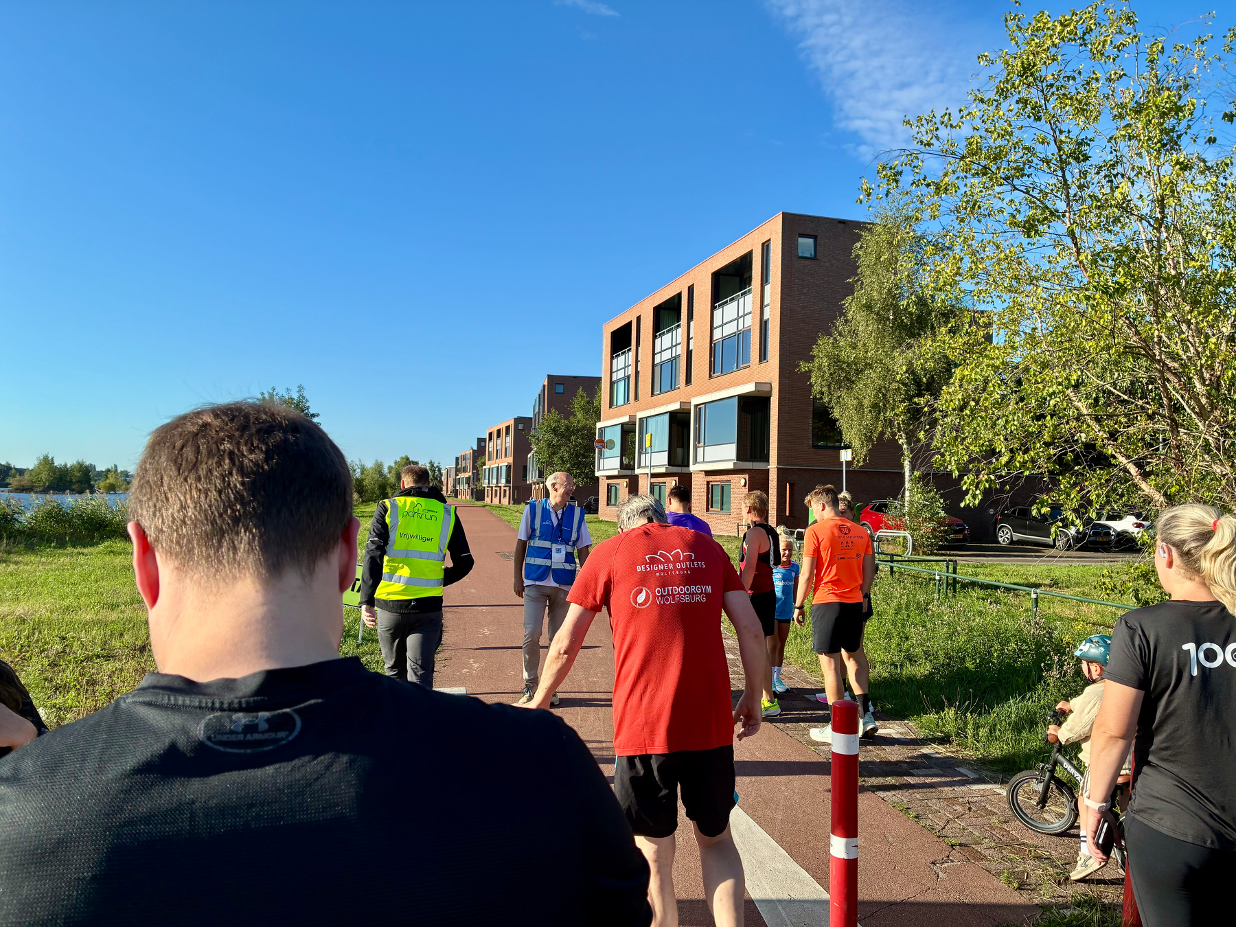 Mensen wandelen en fietsen over een verhard pad naast moderne gebouwen onder een heldere blauwe lucht.