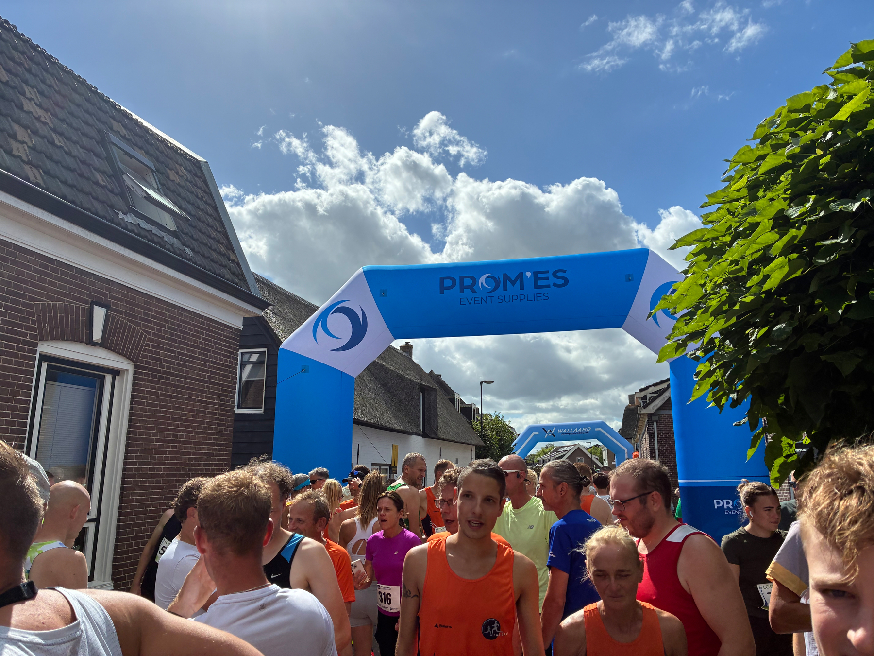 Een groep hardlopers en toeschouwers verzamelt zich onder blauwe opblaasbare bogen met het label PROMES EVENT SUPPLIES op een zonnige dag in een woonwijk.