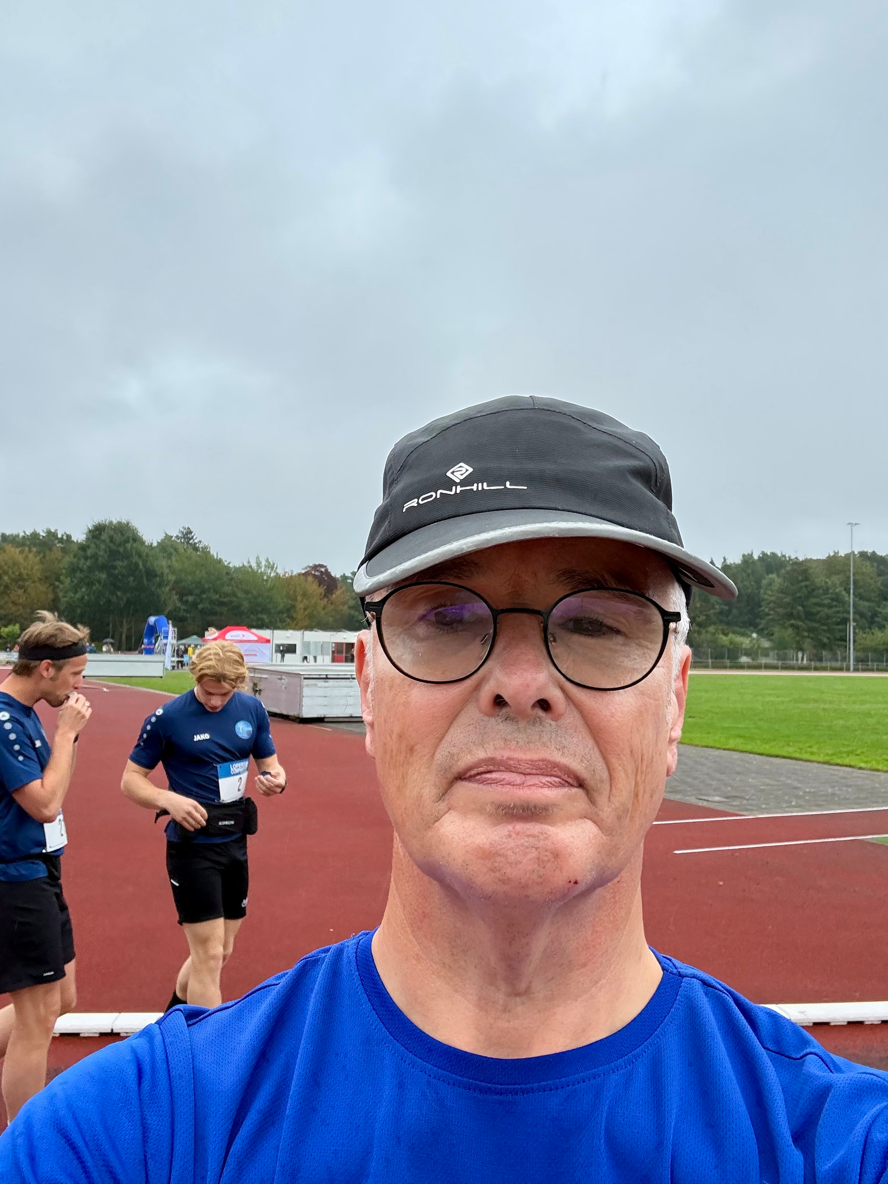 Een man met een bril en een pet maakt een selfie op een hardloopbaan met twee hardlopers in blauwe sportkleding op de achtergrond.