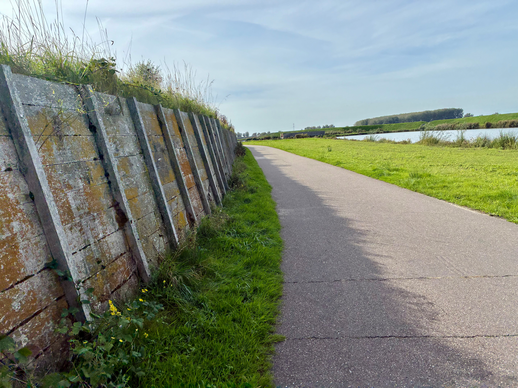Een verhard pad loopt langs een gracht met aan de ene kant een slachtige, met mos bedekte betonnen dijk en aan de andere kant met gras begroeide velden.