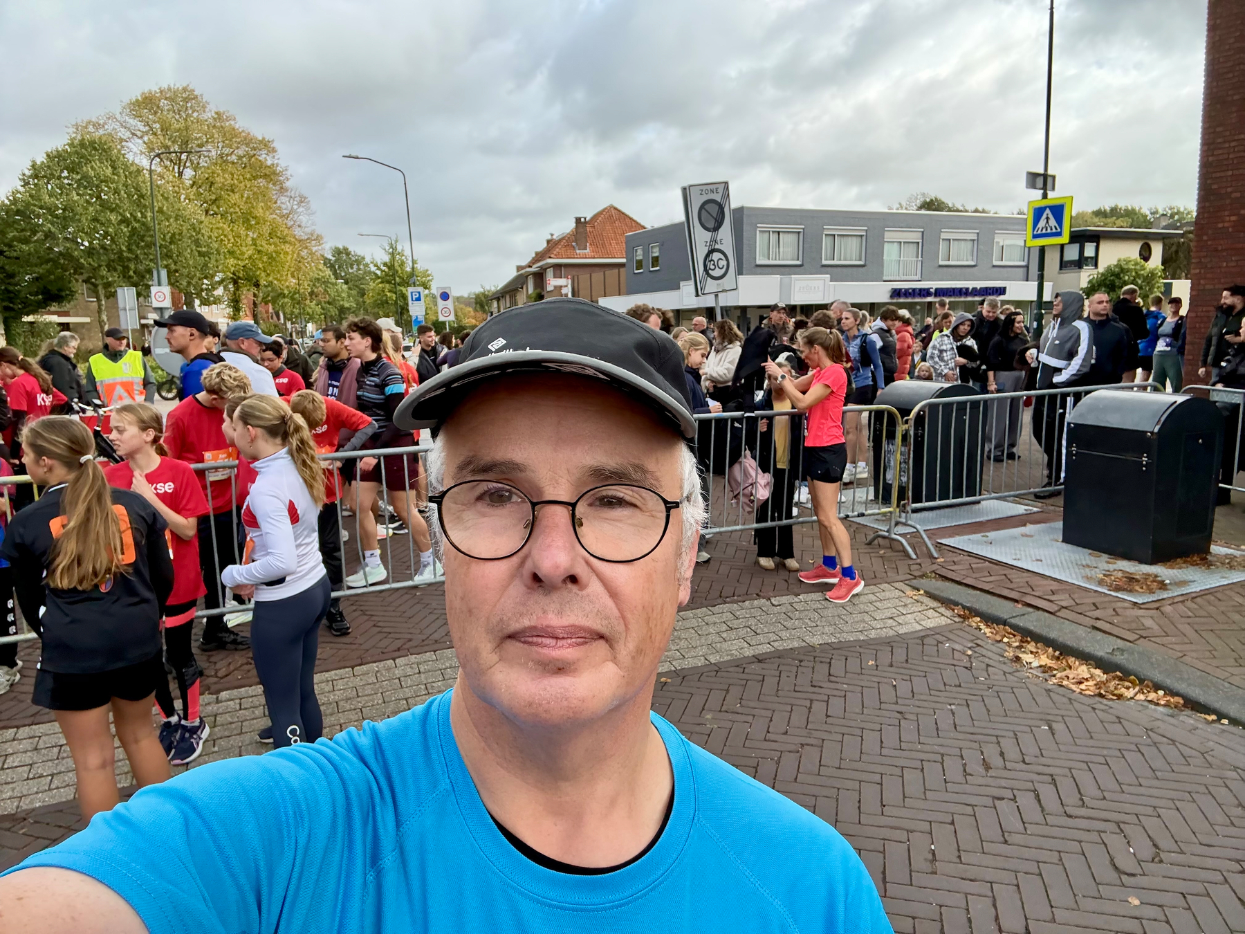 Een man met een bril, een zwarte pet en een blauw shirt maakt een selfie voor een menigte mensen die zich achter een metalen hek op een straat verzamelen.