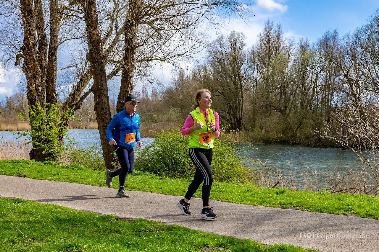 Twee mensen met hardloopkleding joggen langs een pad aan de rivier, omringd door bomen.