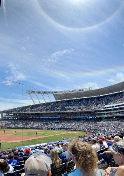 A large crowd is gathered in a baseball stadium under a clear sky with a visible halo around the sun.