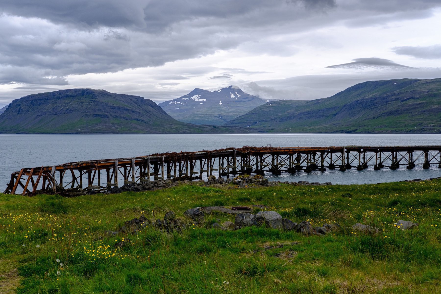 A ruined, rusted harbour with a fjord and mountains in the background