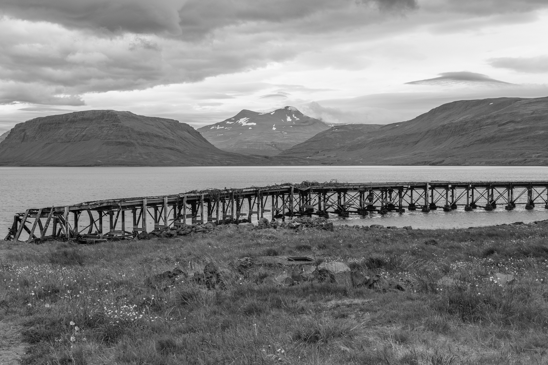 A ruined harbour with Hvalfjörður in the background and a mountain range in the distance. Black and white.
