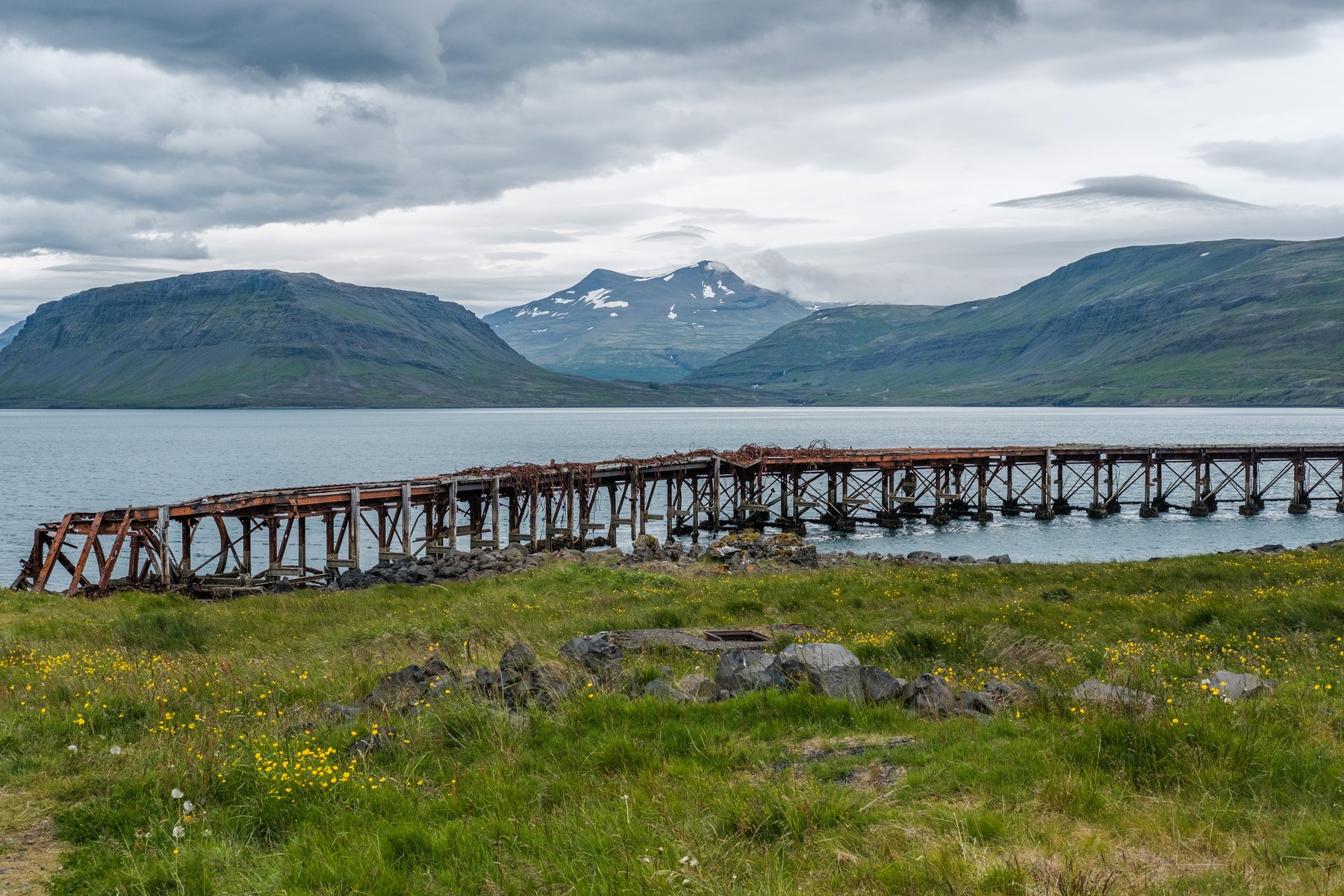 A ruined harbour with Hvalfjörður in the background and a mountain range in the distance. Colour.