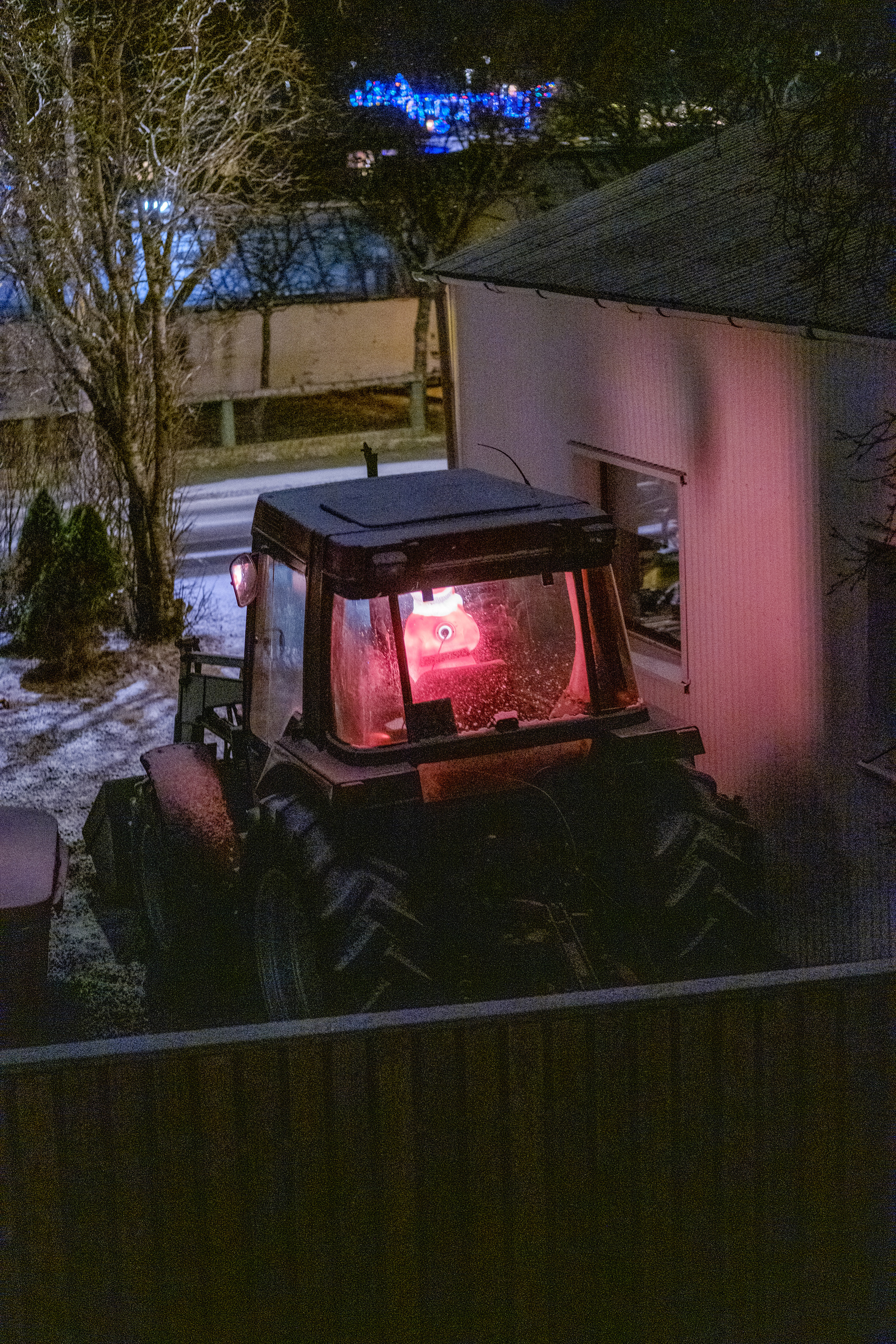 A tractor parked next to a garage. It’s night and the light-up Santa in the tractor’s driver’s seat lights up the wall.