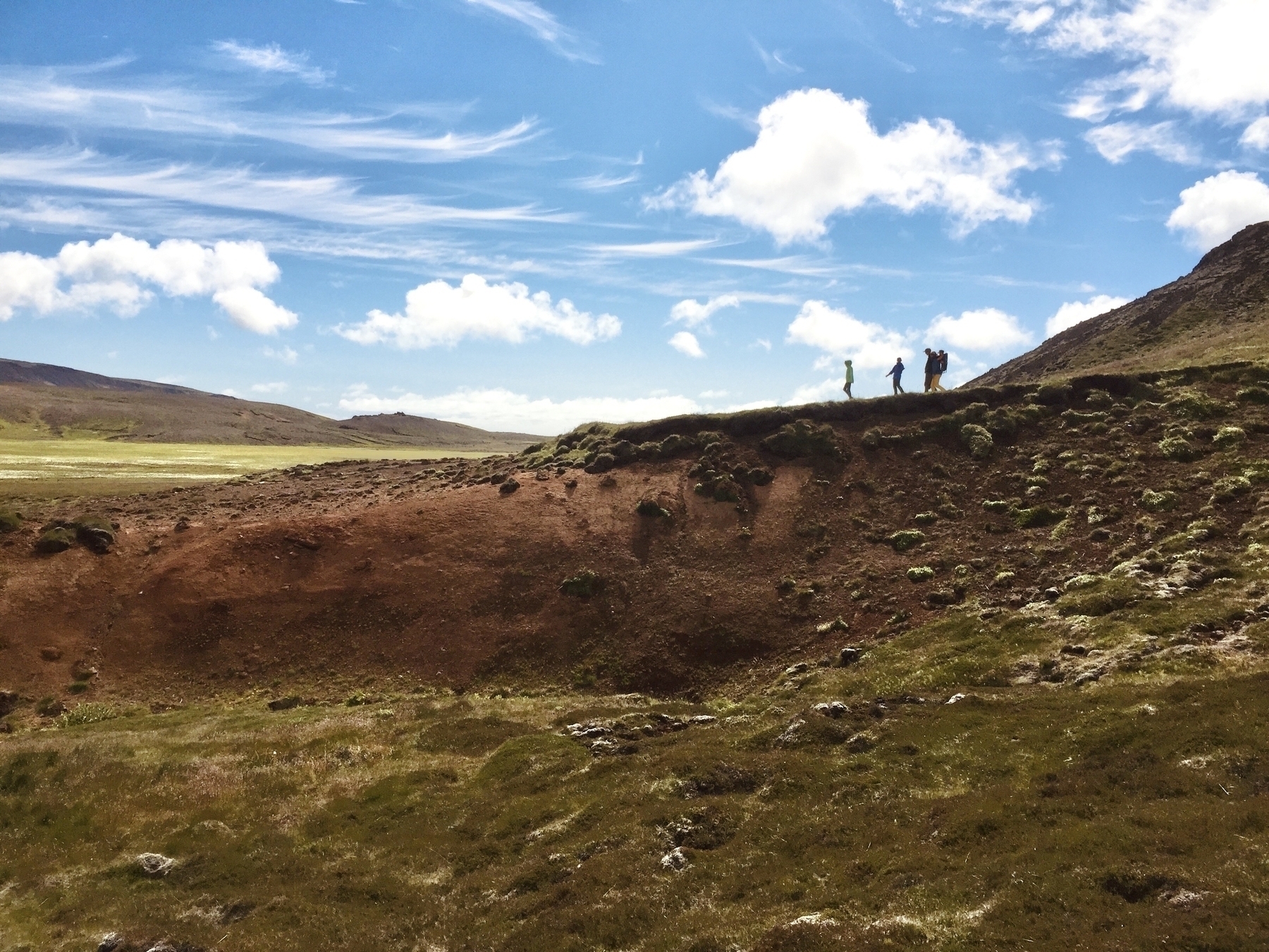 A photo taken several years ago in the geothermal are near the town of Grindavík. A group of tourists are walking along a ridge in the distance.