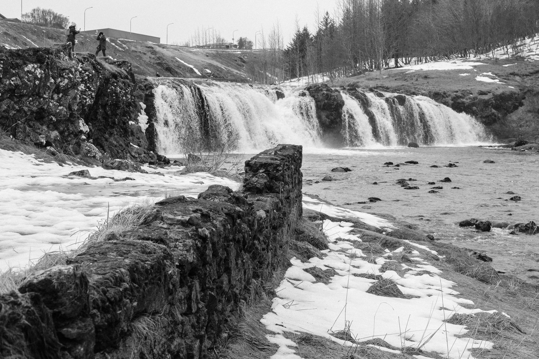 A black and white photo of a small waterfall in the river Varmá. Jutting out from one side of the picture is the ruined wall that's all that's left of the old wool factory