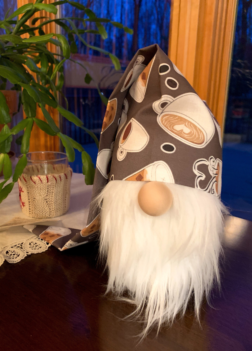 A gnome with a white beard and a coffee-themed hat is sitting on a wooden table next to a plant.