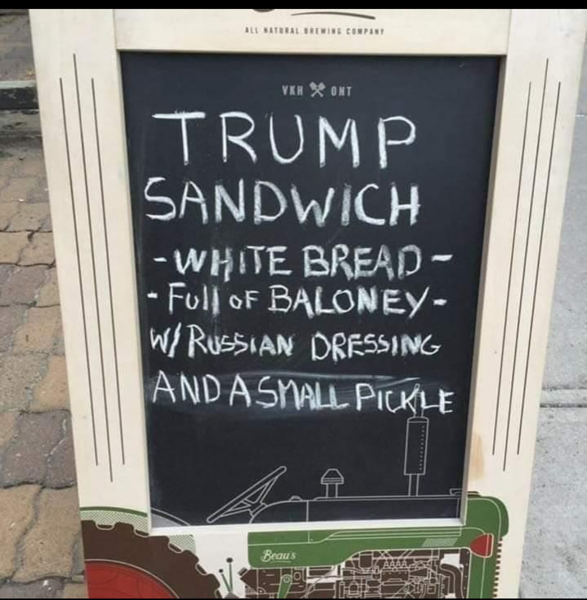An a-frame sign in front of a sandwich shop that reads: Trump sandwich, white bread, full of baloney, with Russian dressing, and a small pickle.