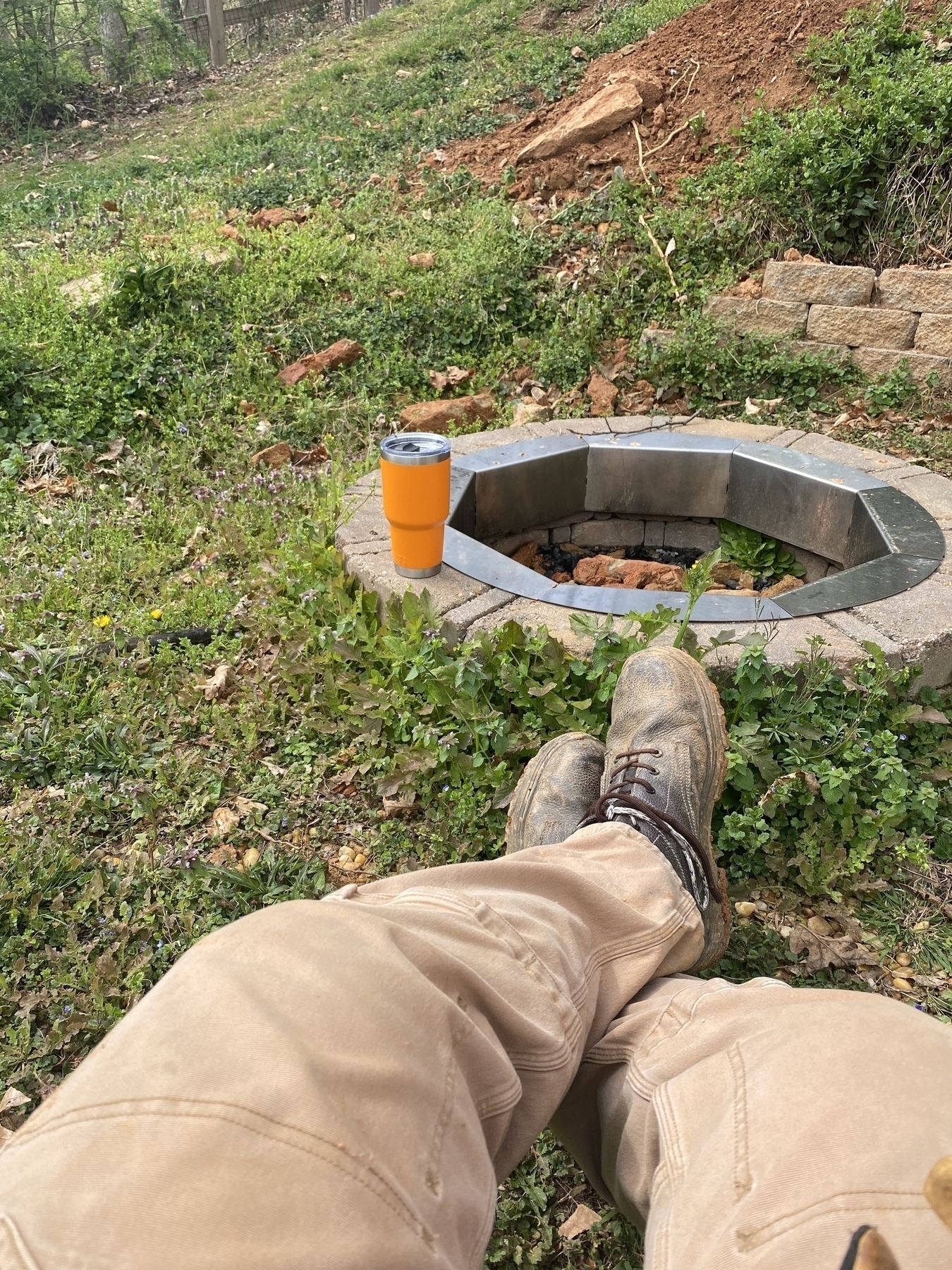 Picture of a man’s pantlegs and boots out in front of him. A fire pit with an orange Yeti on it in the background.