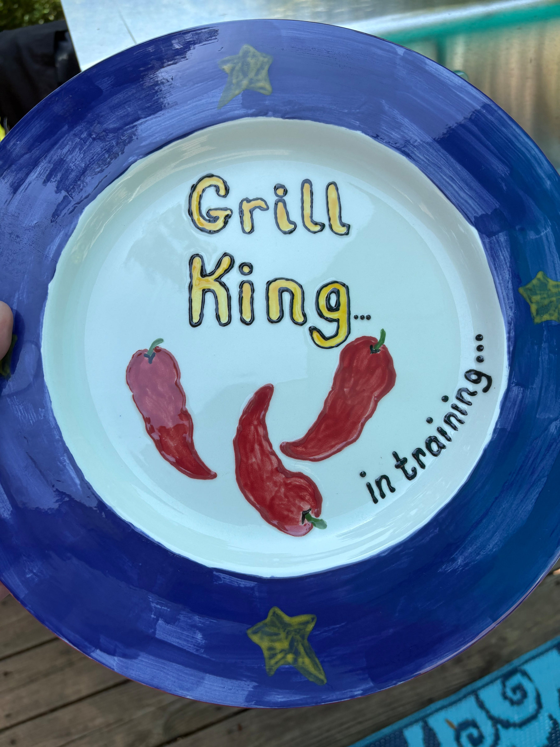 A large plate with a blue outer rim, lettering that reads Grill King in training…, and chili peppers on it.