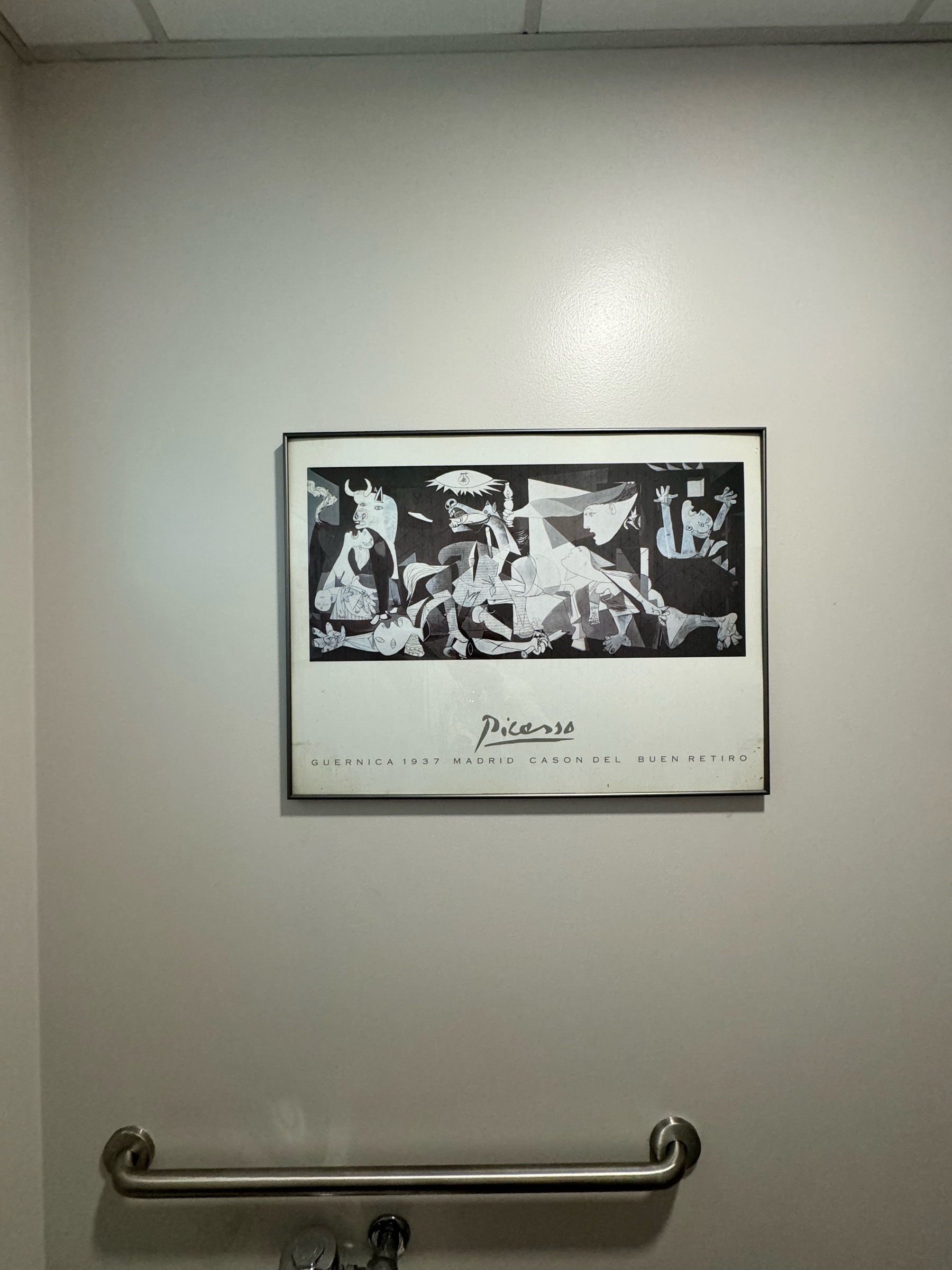 A reproduction of Pablo Picasso's Guernica hangs on the wall above a toilet in a public restroom; you can see the hint of a drop ceiling and an accessibility bar.