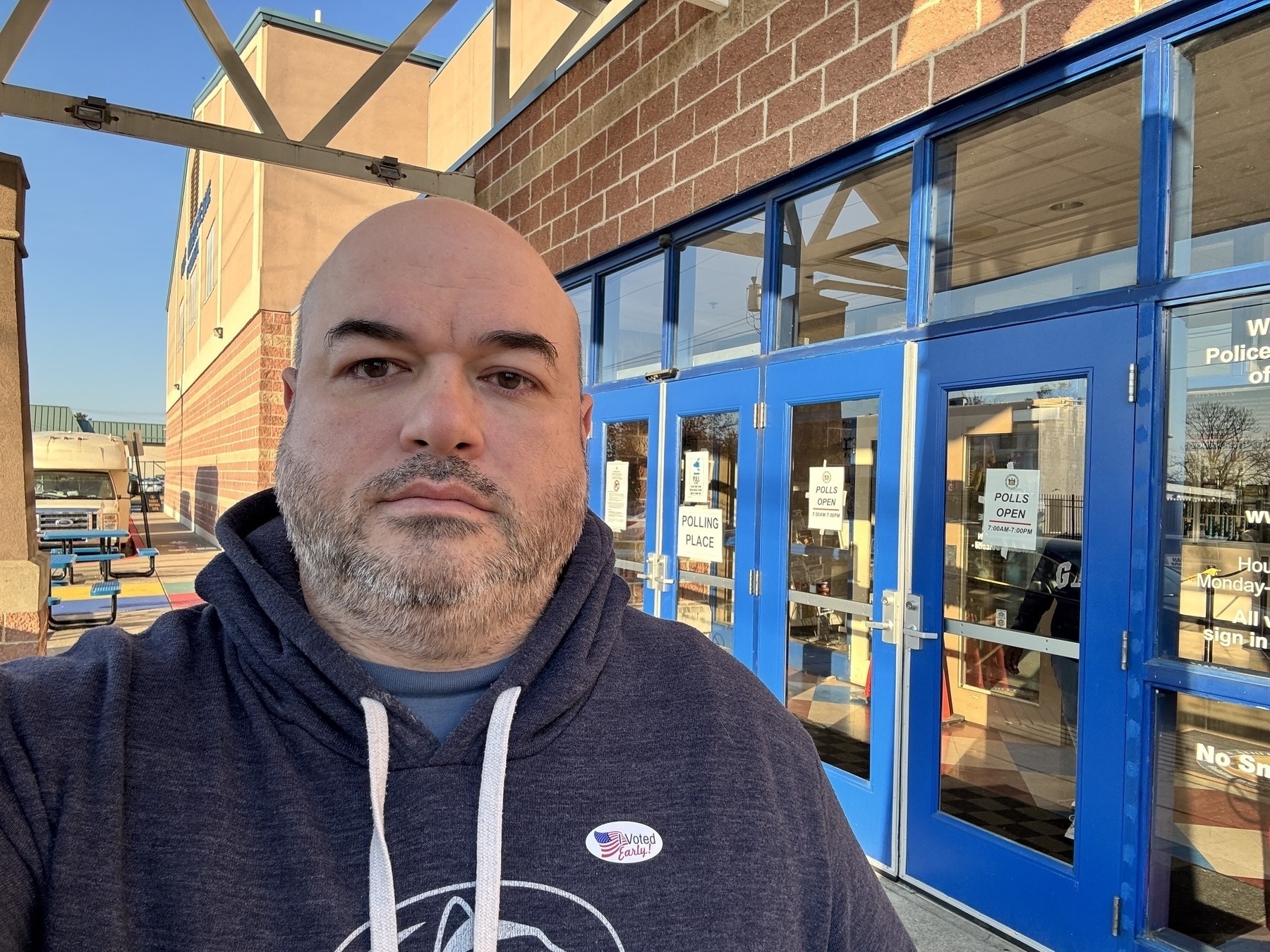 Me, a bald white male with a salt-and-pepper beard, standing in front of the Police Athletic League HQ in Wilmington, Delaware. The outside of the building has signs indicating that it is an early polling place. I have an I Voted Early sticker on my hoodie.