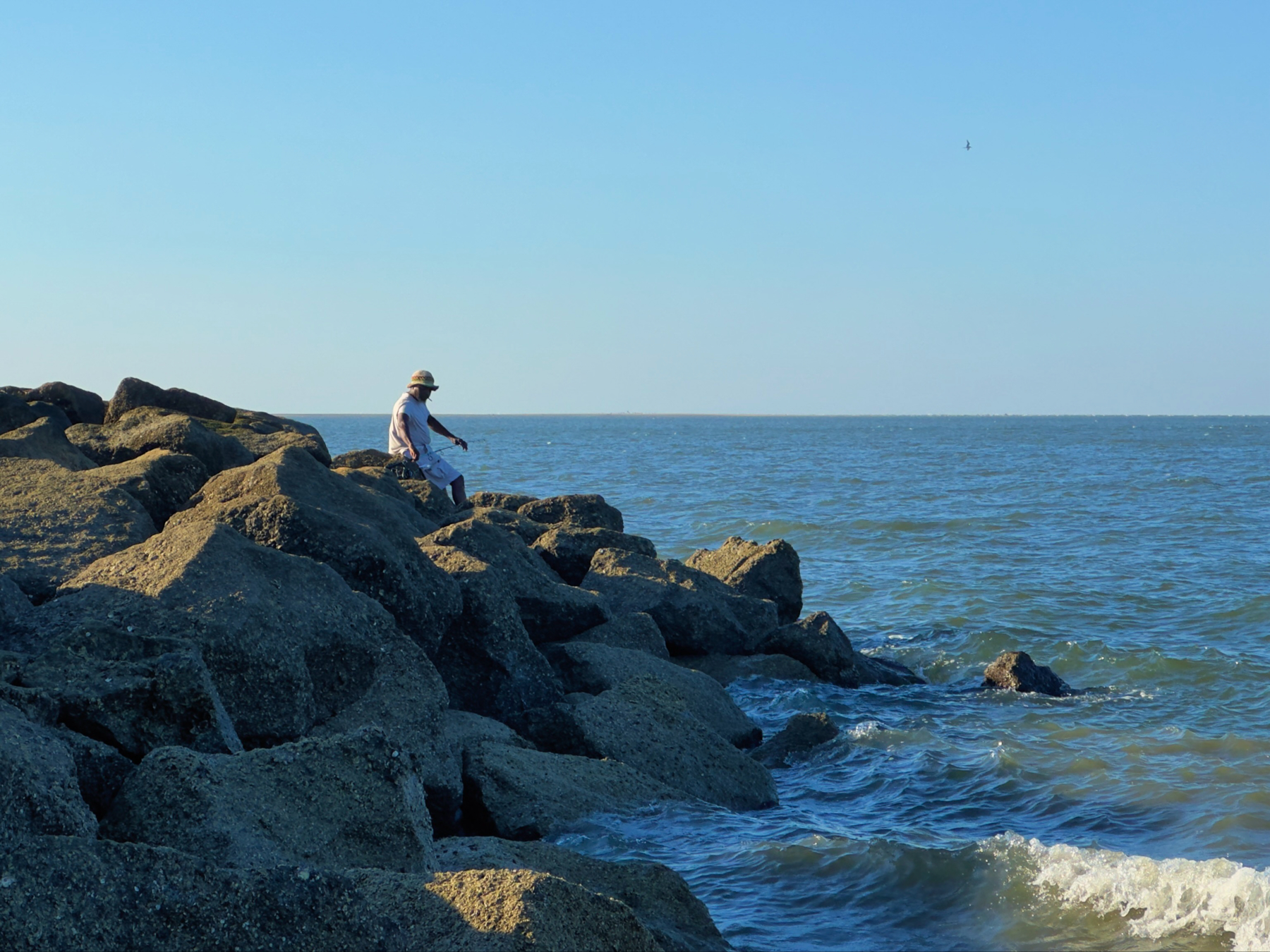 A man sits fishing on a pile of rocks that stretches into the ocean.